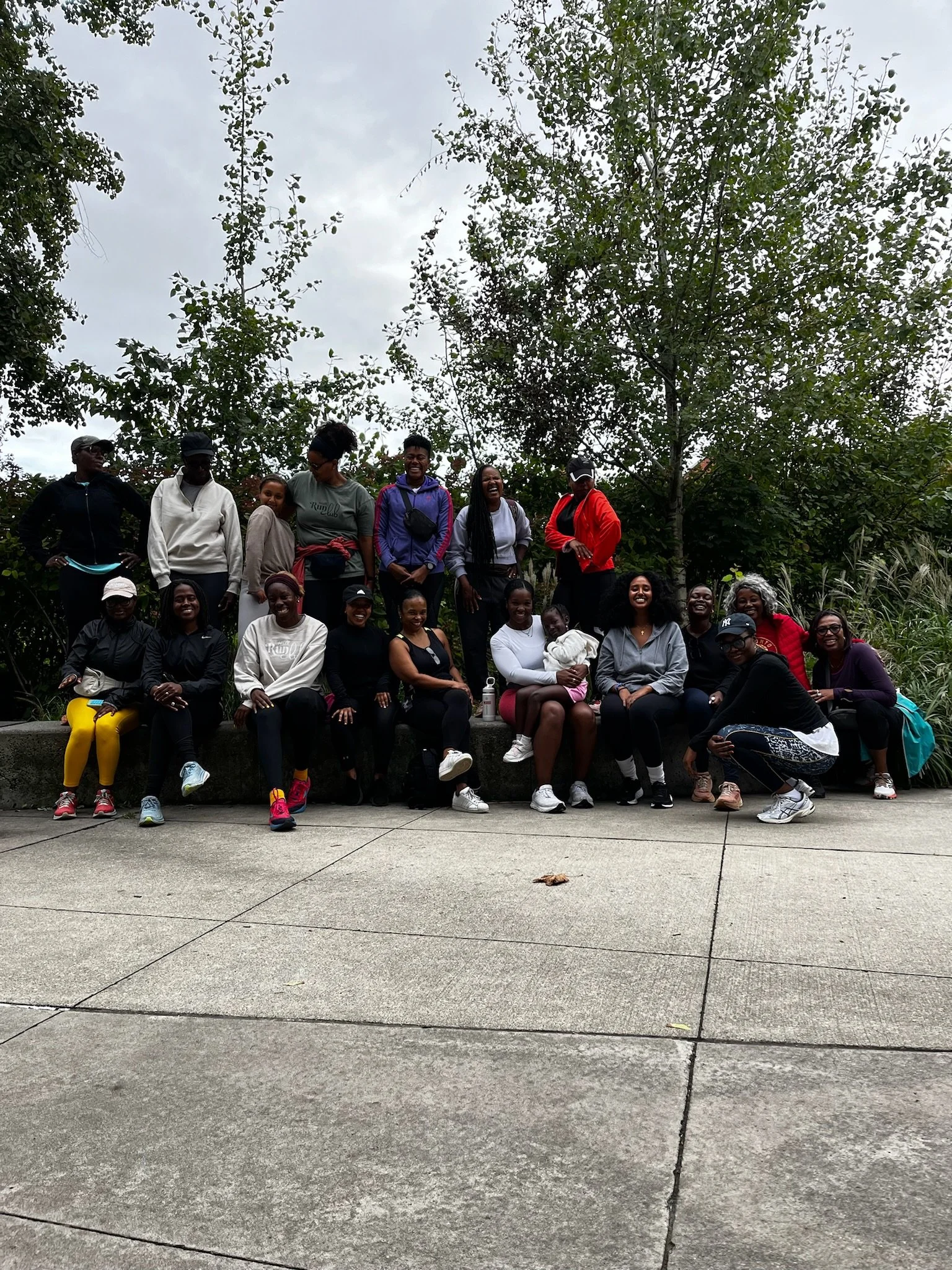 A group of people, mostly women and girls, sitting and standing outdoors on a ledge and pavement, smiling and posing for a photo, with trees and a cloudy sky in the background.