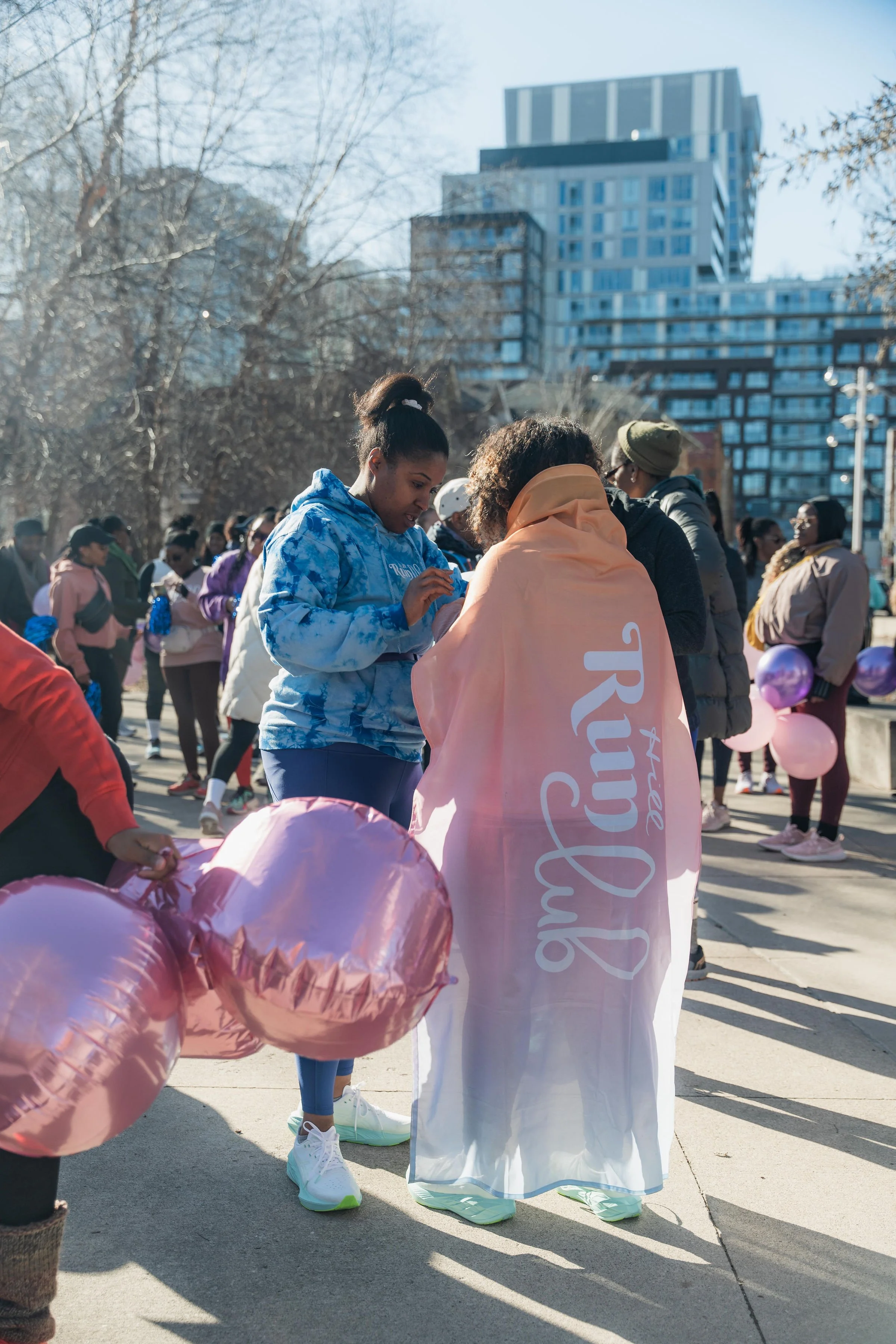 Group of women participating in a race or walk event, with one woman wearing an orange and pink cape that reads 'True Run Club,' holding pink heart-shaped balloons, with other participants in the background in a city park on a sunny day.