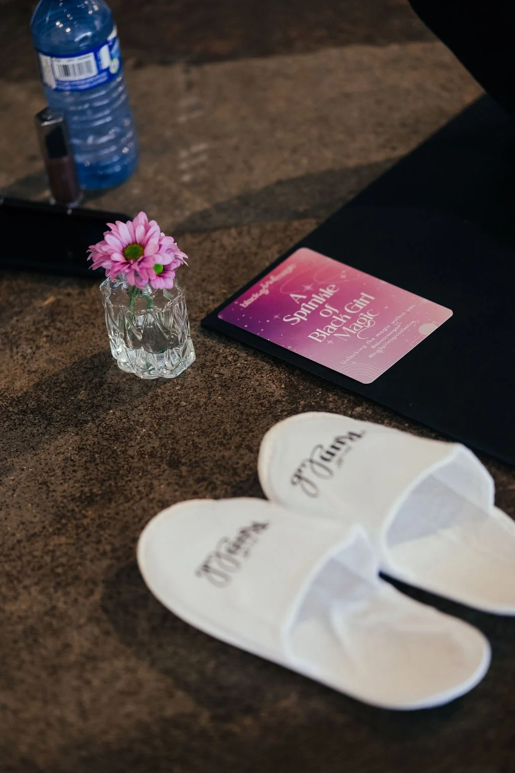 A small pink flower in a clear decorative glass vase placed on a dark brown table, with a water bottle, a dark folder, a pink card titled 'A Sparkle of Black Girl Magic,' and a pair of white slippers with 'MUDAGE' written on them.