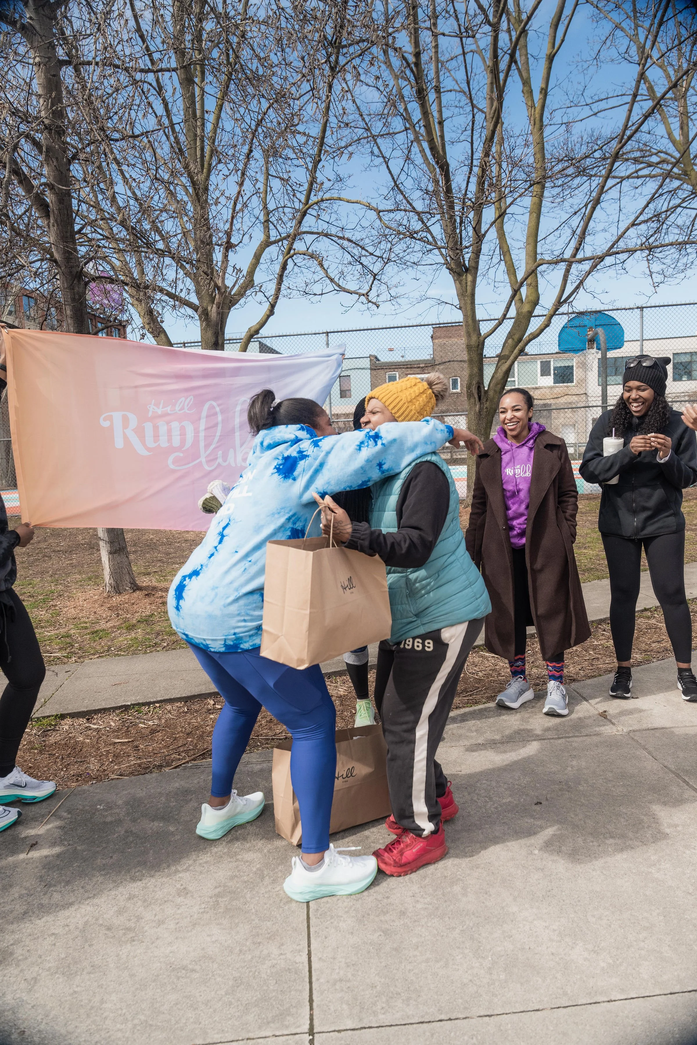 Group of women celebrating outdoors, with two women hugging and one woman holding a paper bag. They are standing near a banner that reads 'Hill Run Club' with leafless trees and a fence in the background, suggesting a park setting on a clear, sunny d