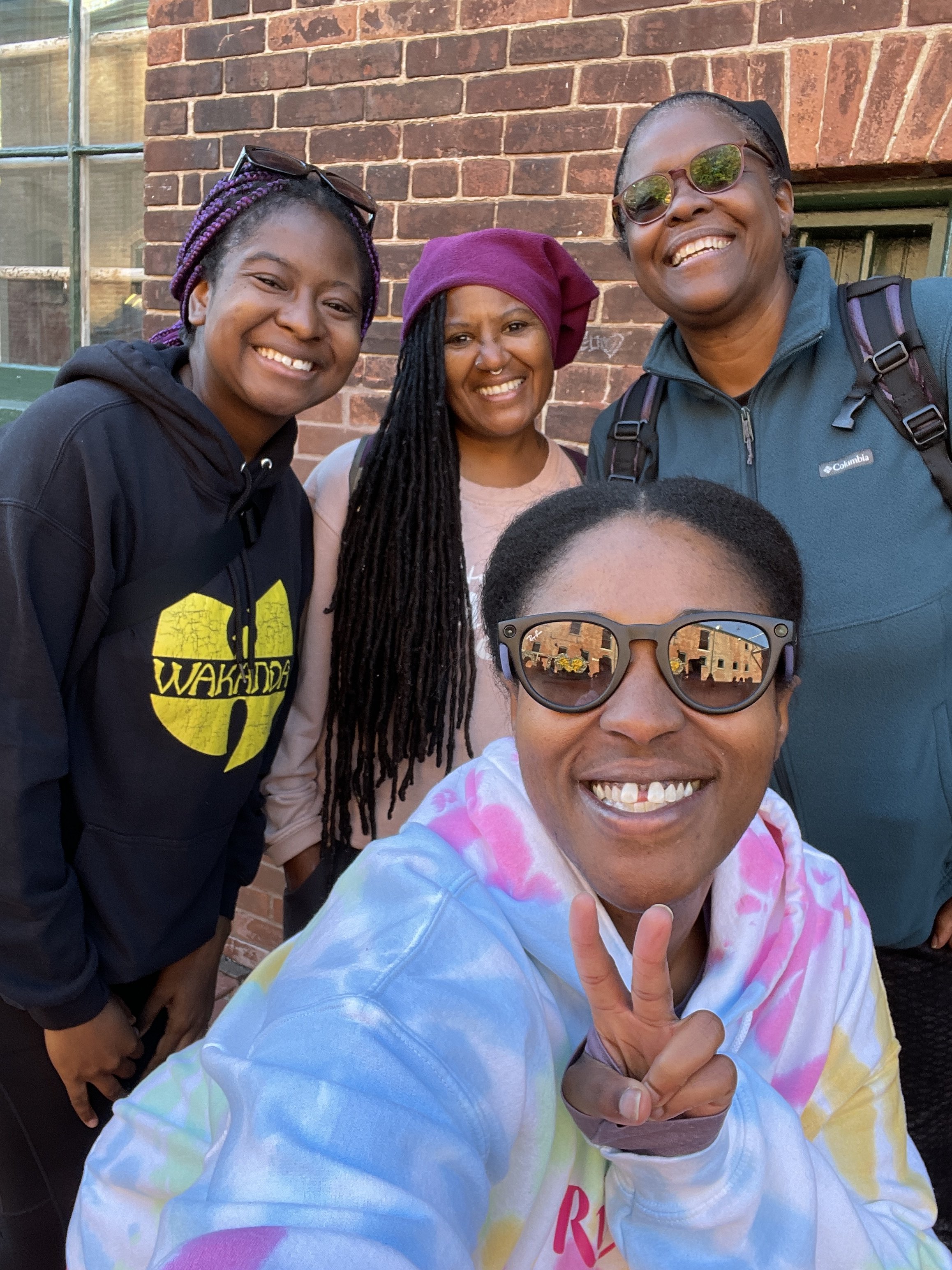 Group of five women smiling and taking a selfie outside against a brick wall, with one woman in front wearing sunglasses and showing a peace sign.
