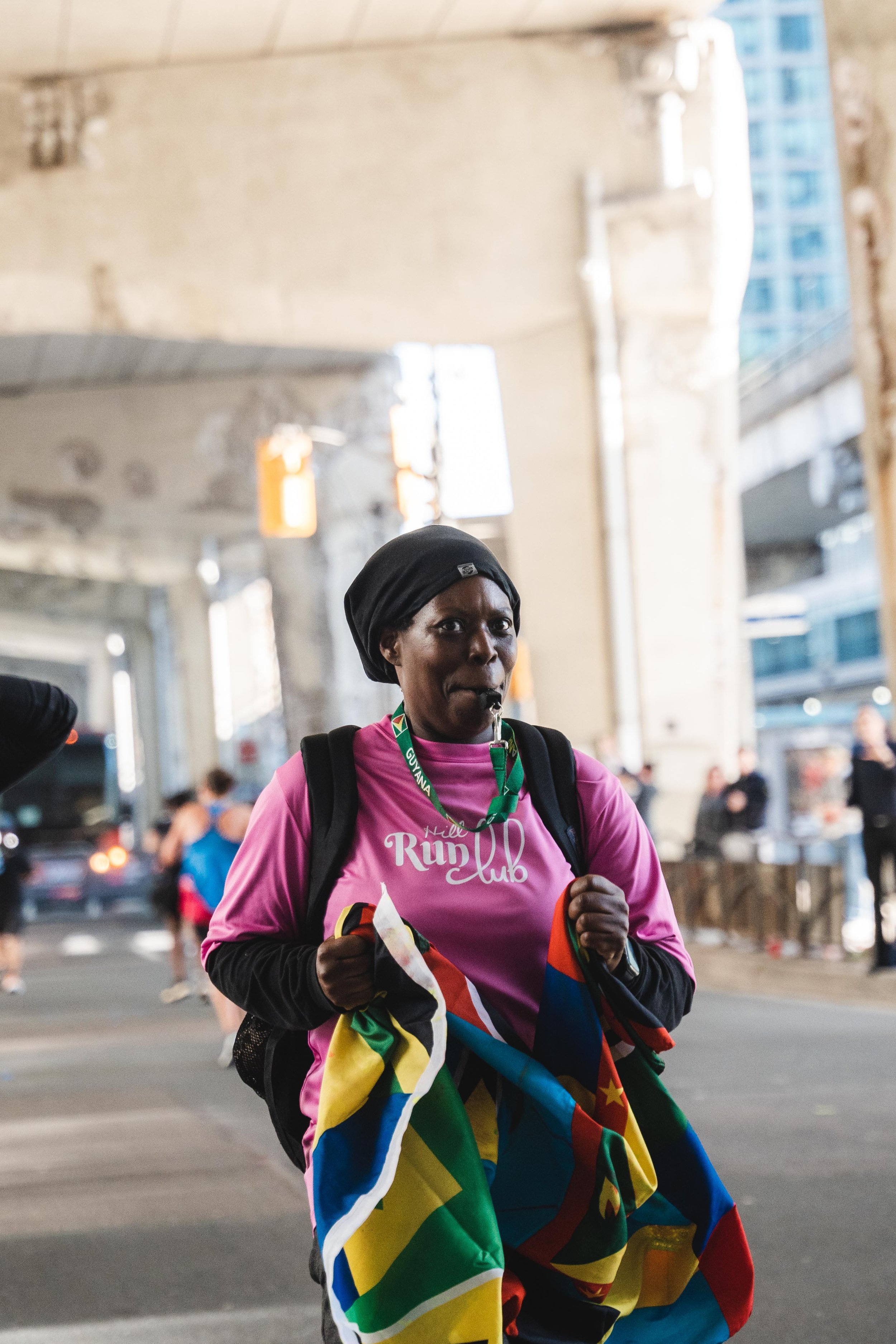 A woman wearing a pink shirt with the text 'Hill Run Club', black head covering, and a backpack holding a colorful flag, running under an overpass with other runners and spectators in the background.