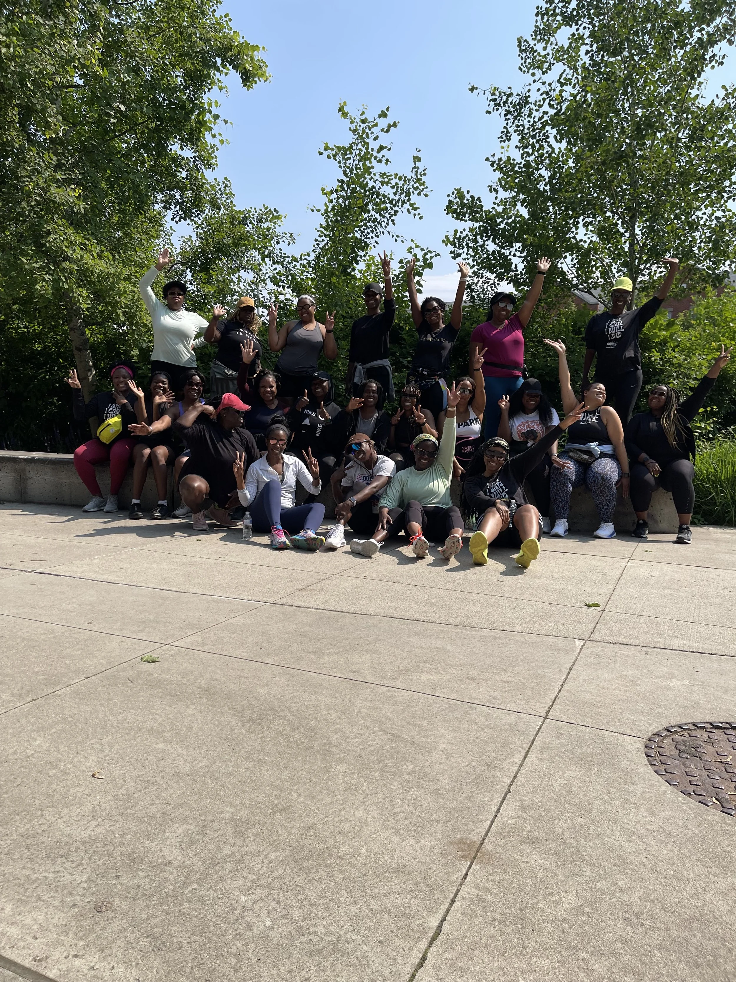 Group of diverse people posing outdoors, some standing and some sitting, cheerful, with trees in the background and sunny weather.