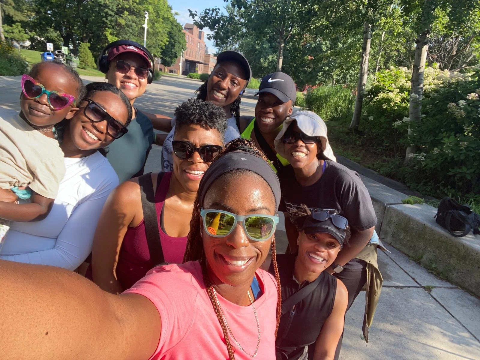 Group of nine women and a girl taking a selfie outdoors on a sunny day, smiling and wearing casual clothing and sunglasses, with trees and a sidewalk in the background.