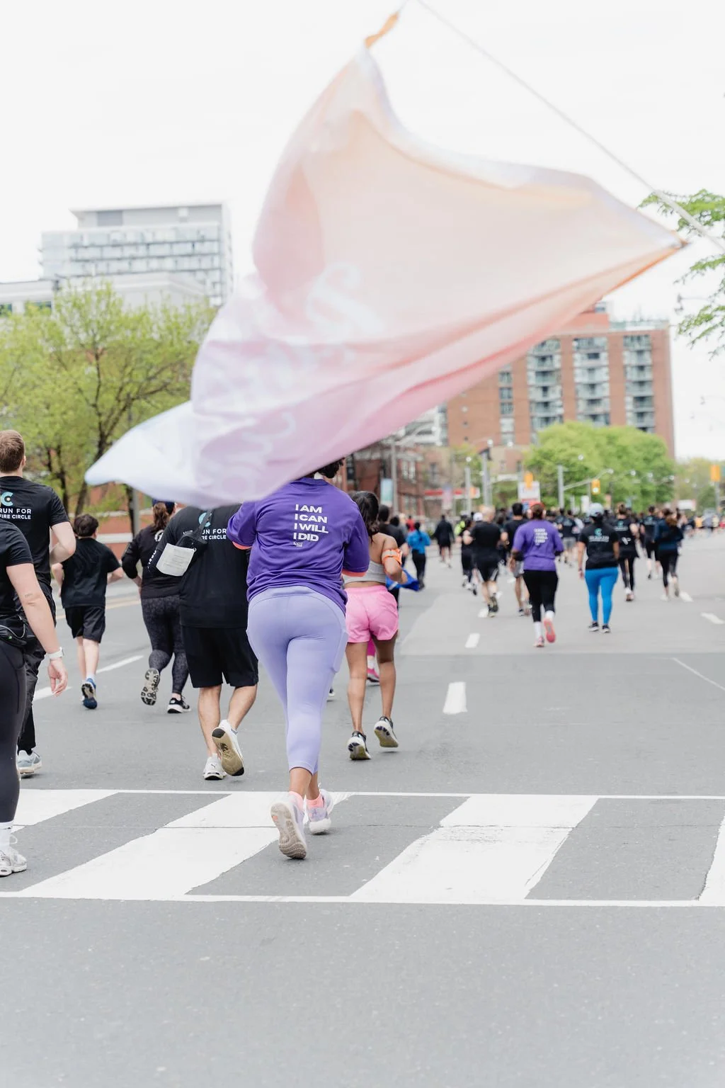 People participating in a marathon or organized run on a city street, with some wearing running clothes and a large pink flag being waved.