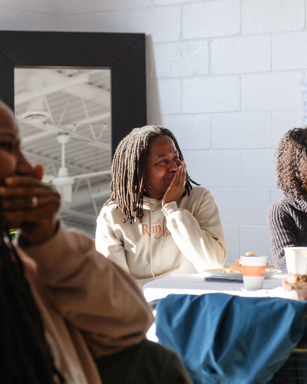 A woman with dreadlocks wearing a beige hoodie, sitting at a table, covering her mouth with her hand, smiling or laughing.