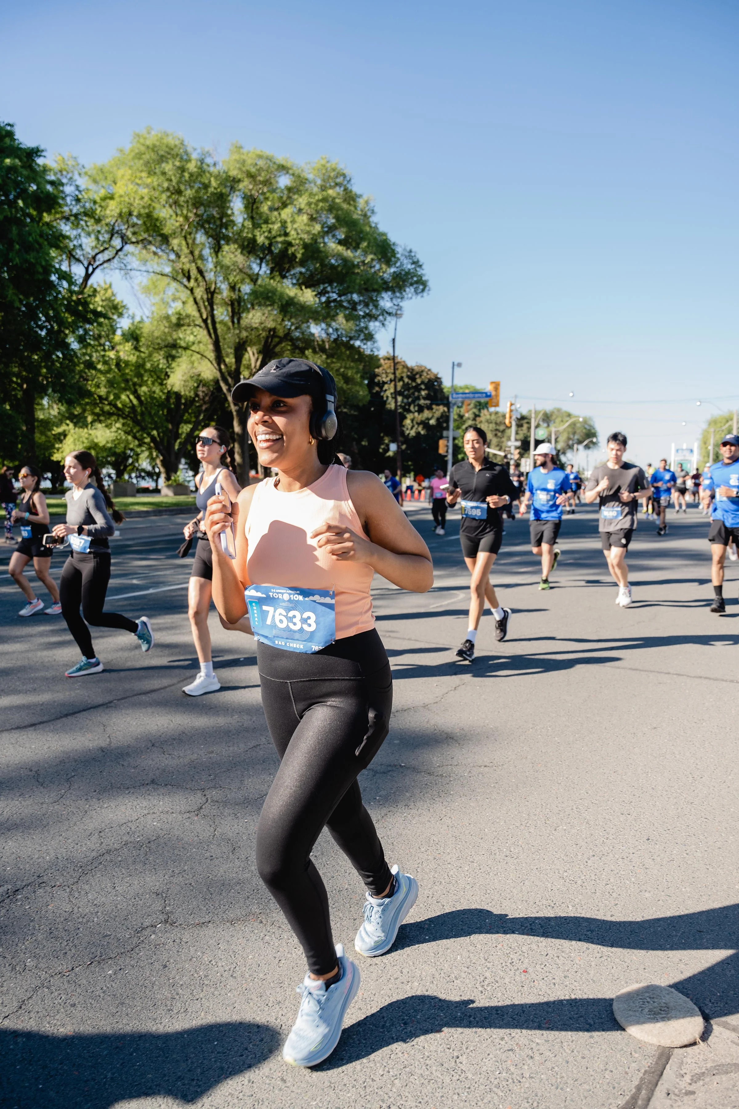 A woman running in a marathon, wearing a light pink tank top, black leggings, a black cap, and headphones, running on a city street with green trees and other runners in the background on a sunny day.