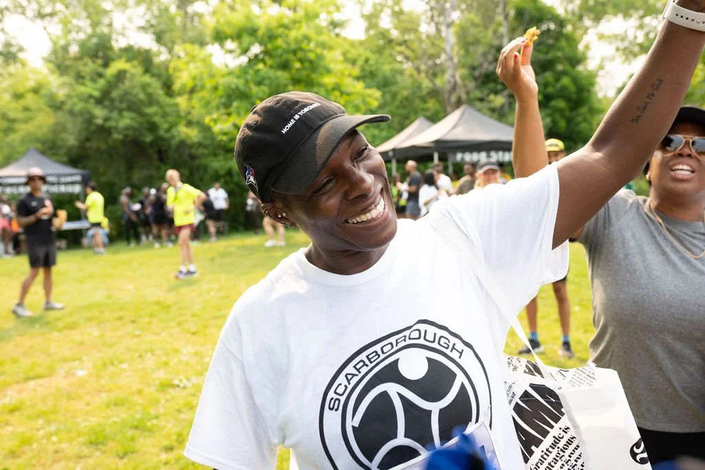 Smiling woman with a black cap and white t-shirt, outdoors at a community event with people, tents, and trees in the background.
