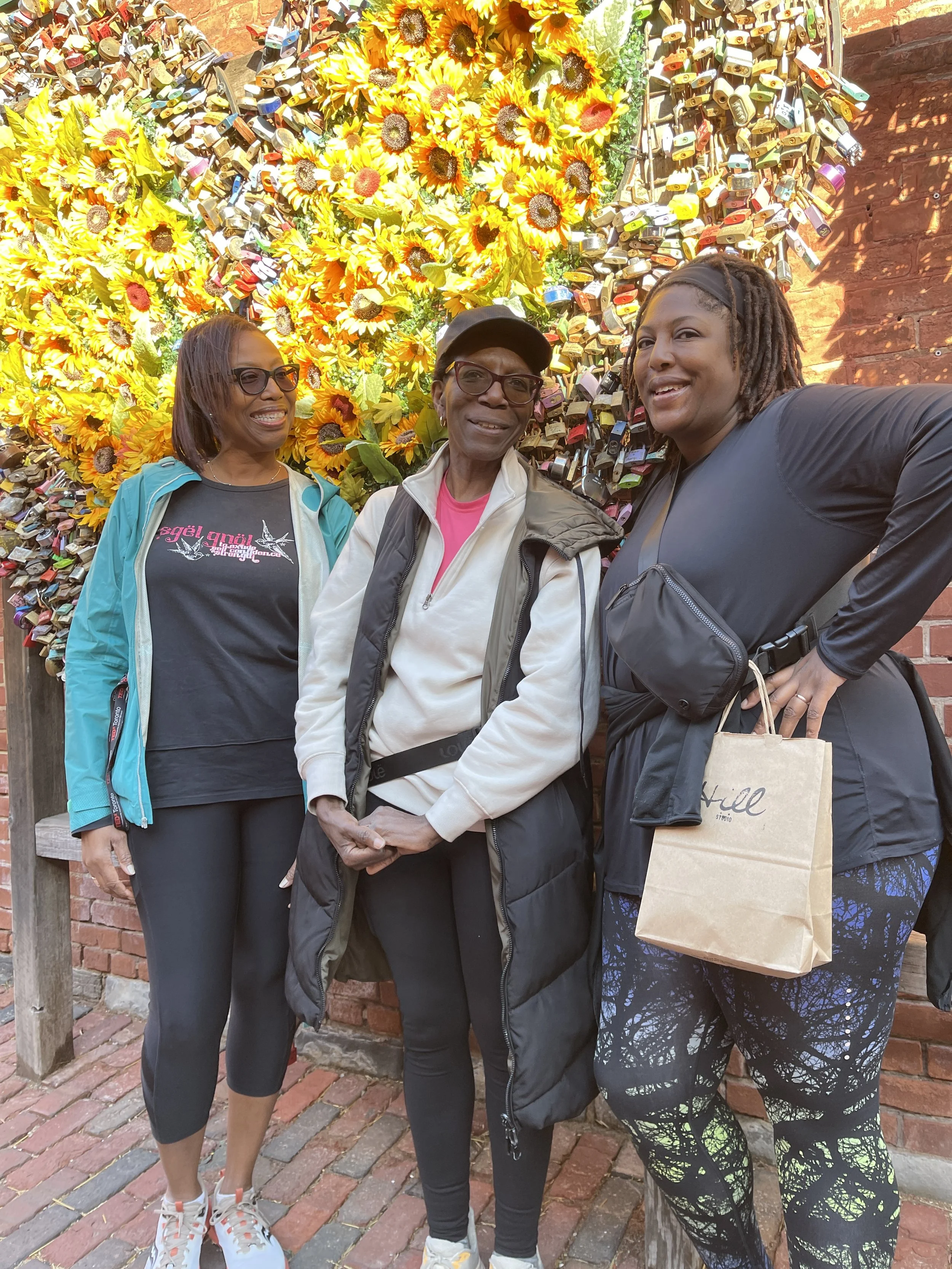 Three women smiling and taking a photo in front of a wall decorated with sunflowers and numerous love locks.
