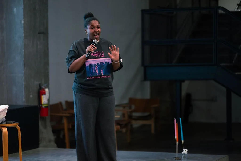 A woman speaks into a microphone, standing on a stage with a gray wall behind her. She wears a black T-shirt and gray pants, and has her hair in a bun. There are candles and chairs in the background.