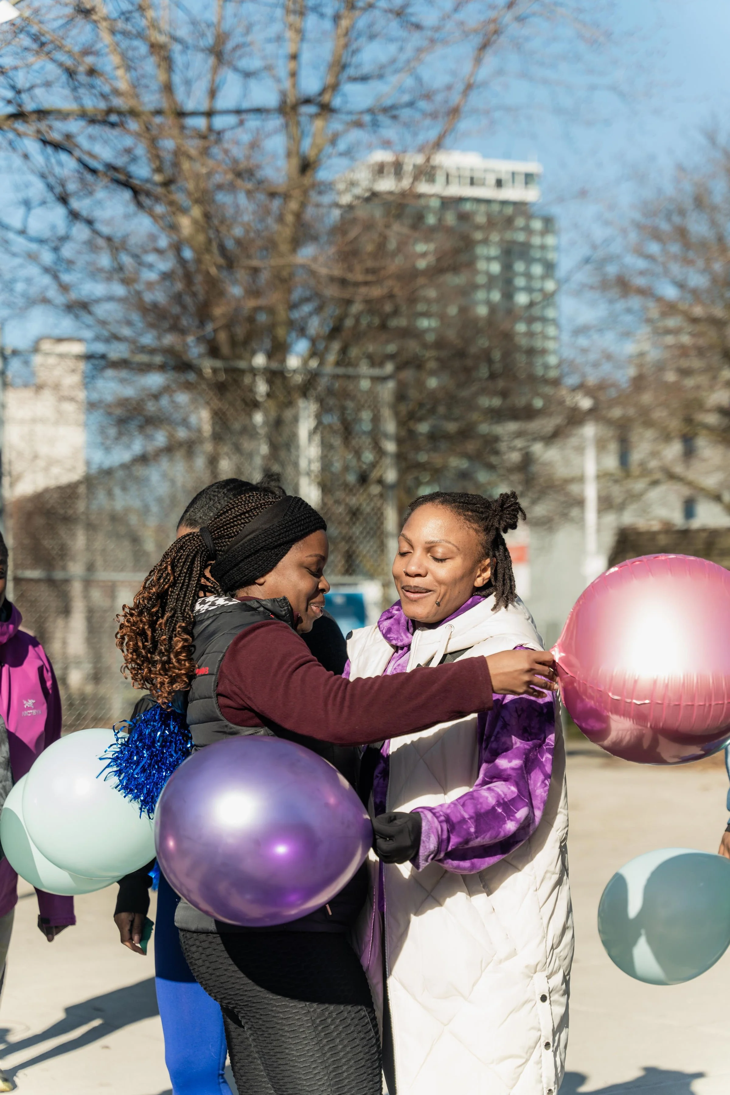 Two women exchange a hug during a celebration, holding balloons, with additional people in the background on a sunny day in an outdoor urban park.