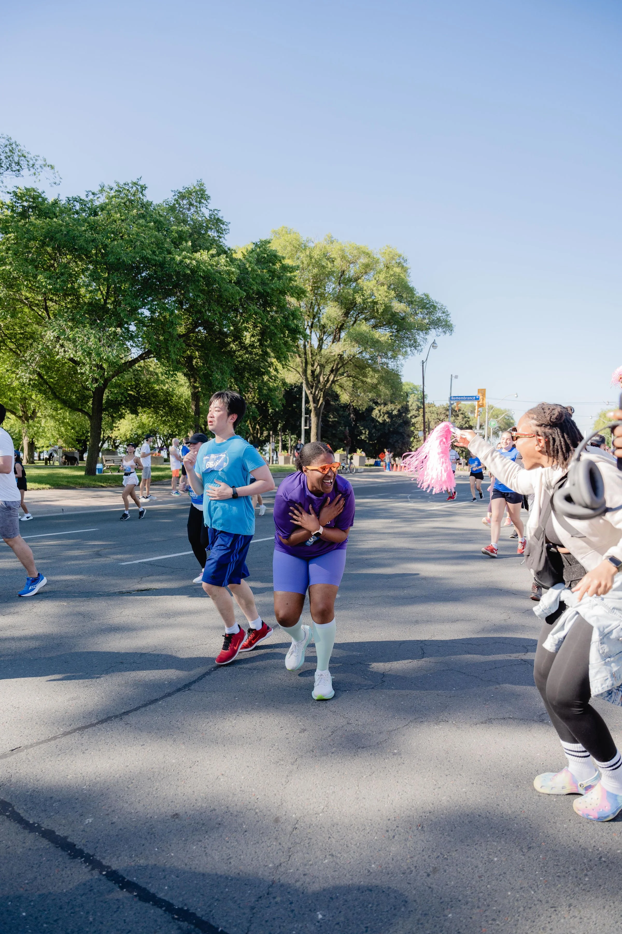 People participating in a fun run or race event on a sunny day, with a woman smiling and crossing the finish line or an obstacle, surrounded by greenery and other runners.