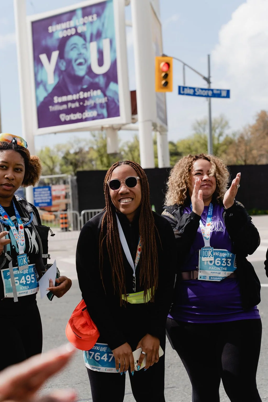 Three women at a running event, posing for a photo, with race bibs, one making a hand gesture, in front of a traffic light and a street sign.