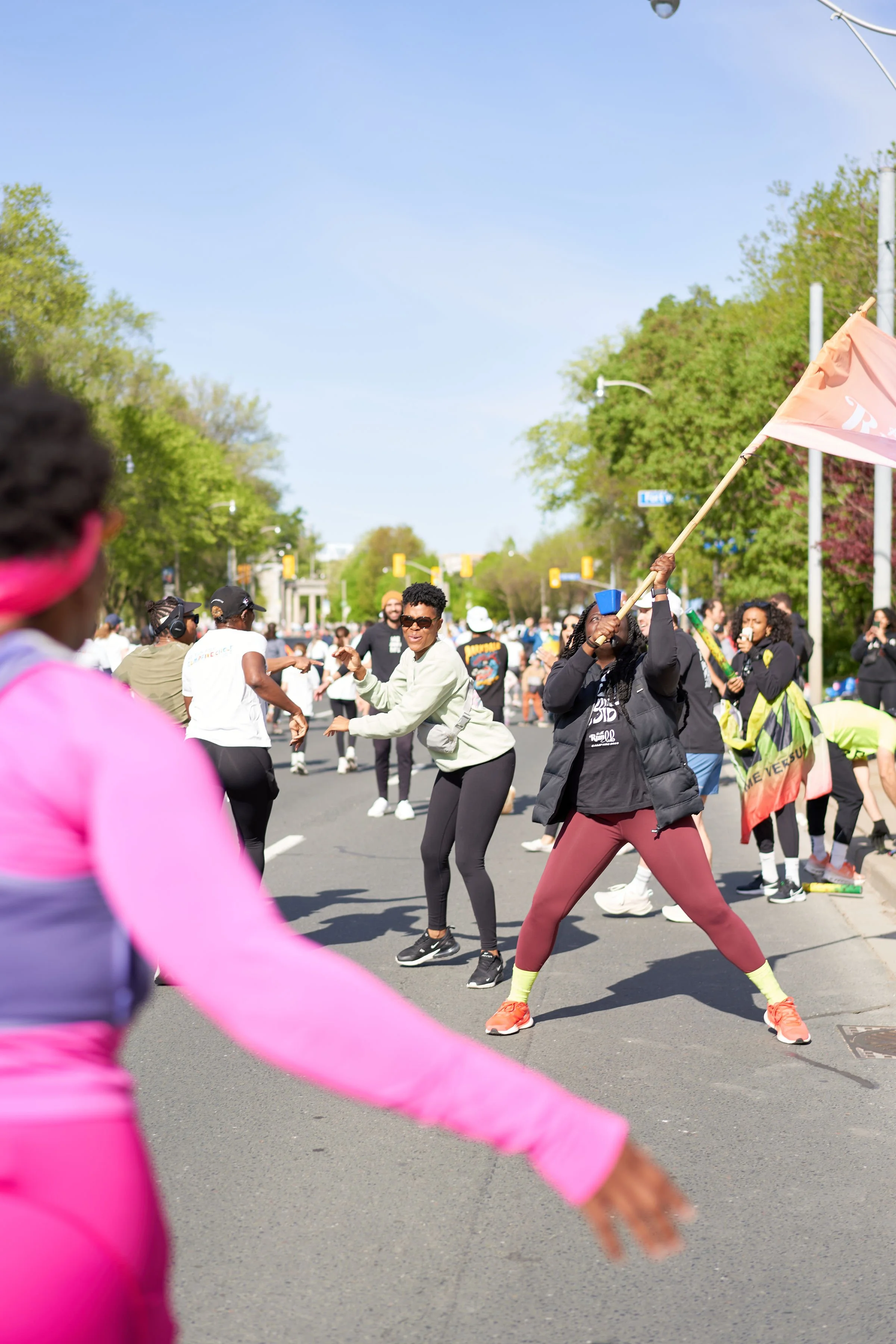 People dancing and celebrating in a parade on a sunny day, with trees in the background and colorful clothing.