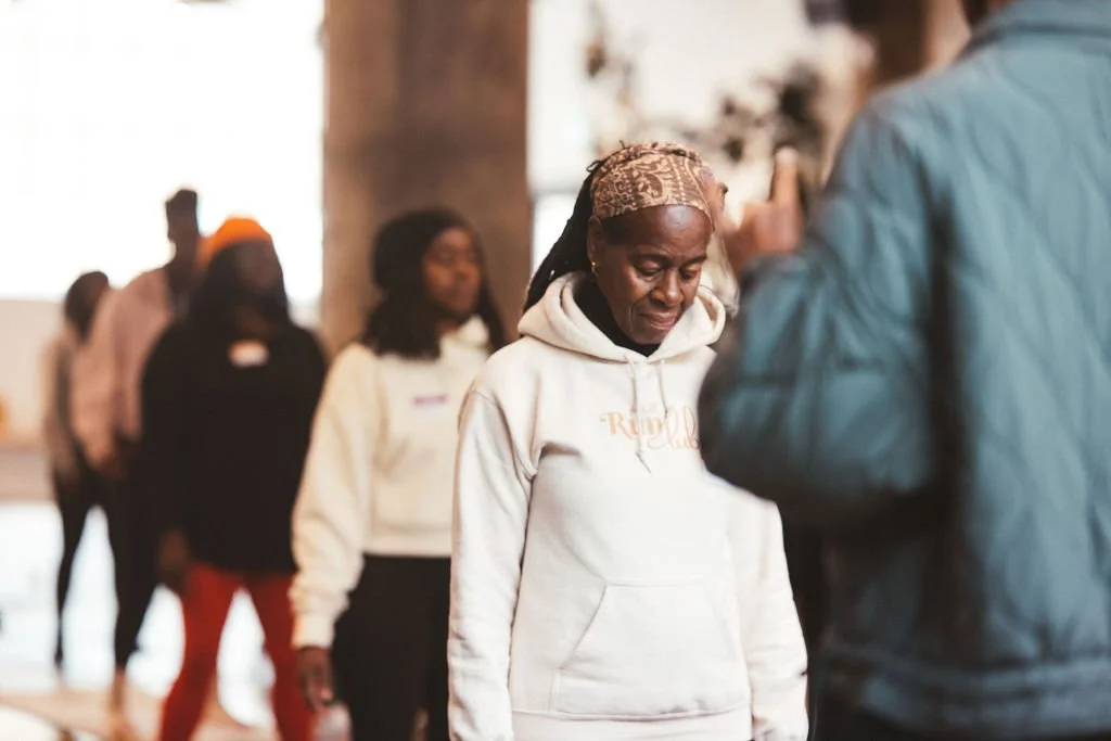 A group of people standing in line, with a woman in the foreground looking down, wearing a white hoodie and a patterned headscarf.