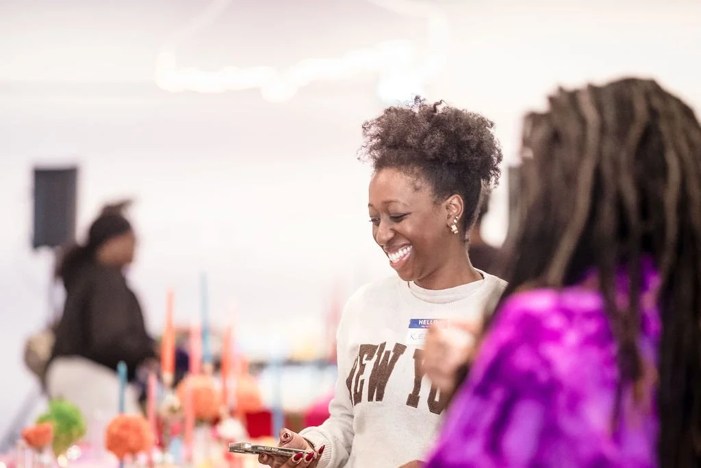 Smiling woman with curly hair looking at her phone at a celebration with pink, blue, and orange decorations in the background.