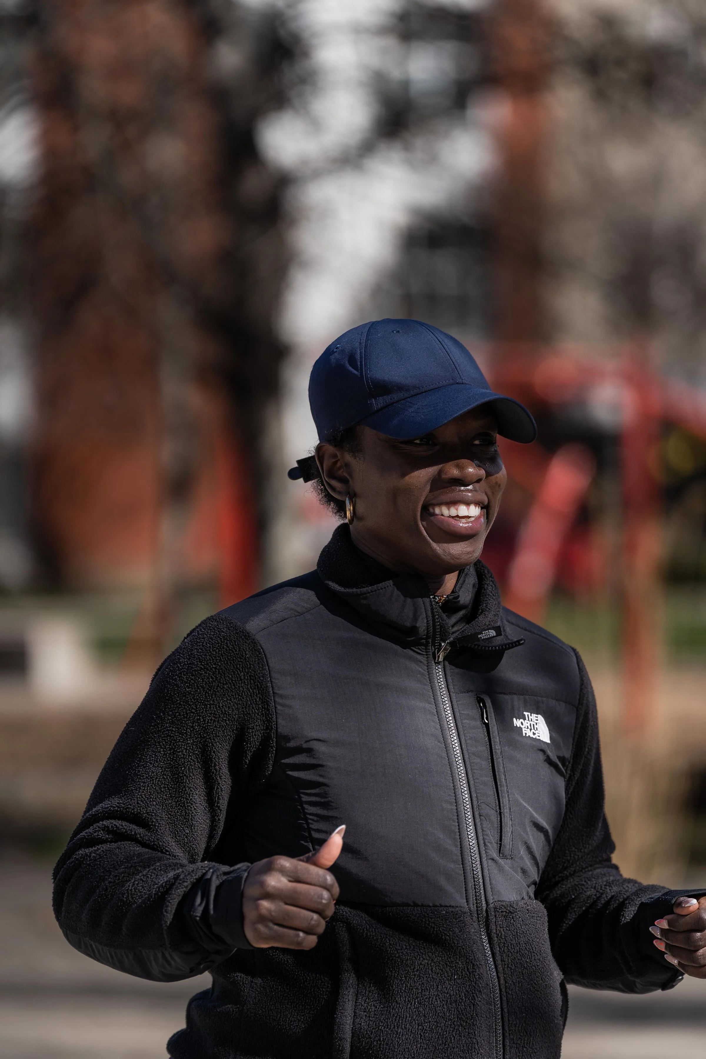 A woman running outdoors, wearing a blue cap and a black jacket, smiling.
