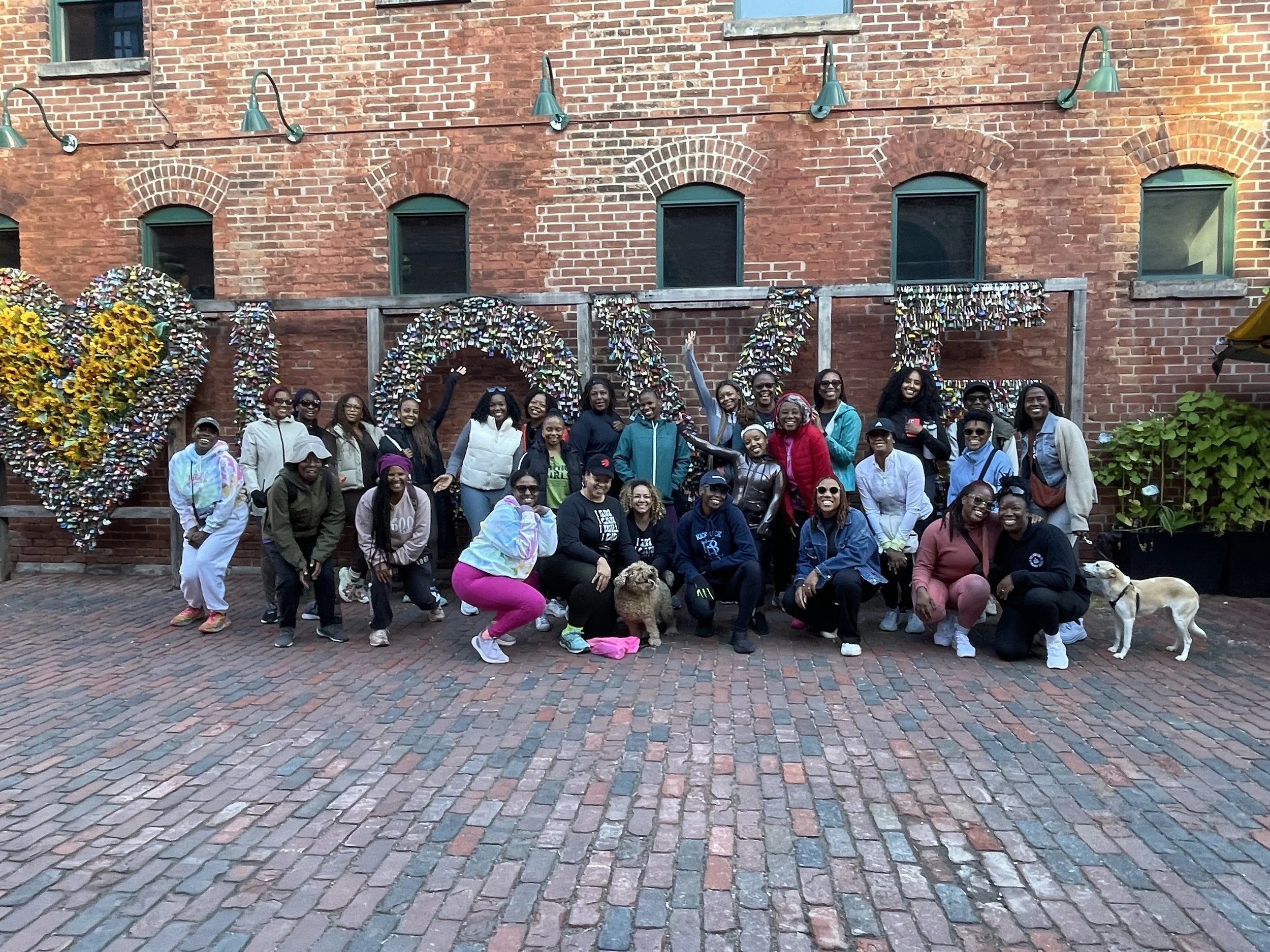 Group of diverse women and children posing outdoors in front of a brick wall with love-shaped decorations made of flowers and padlocks. Two dogs are also present.