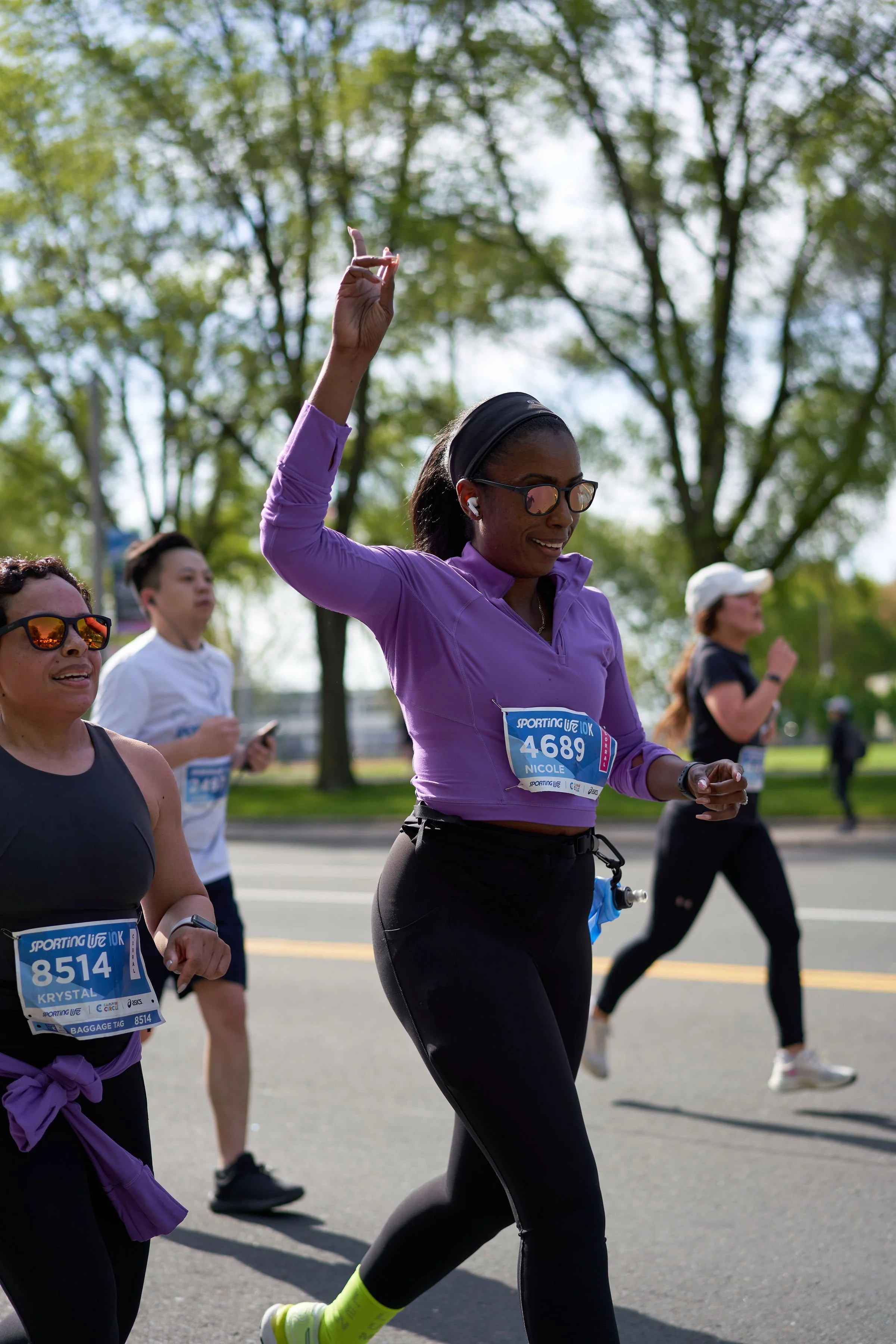 A group of marathon runners participating in a race, with trees in the background. The woman in the foreground is wearing a purple athletic top, black leggings, sunglasses, and has her hand raised as if waving or signaling. She has a race bib number 