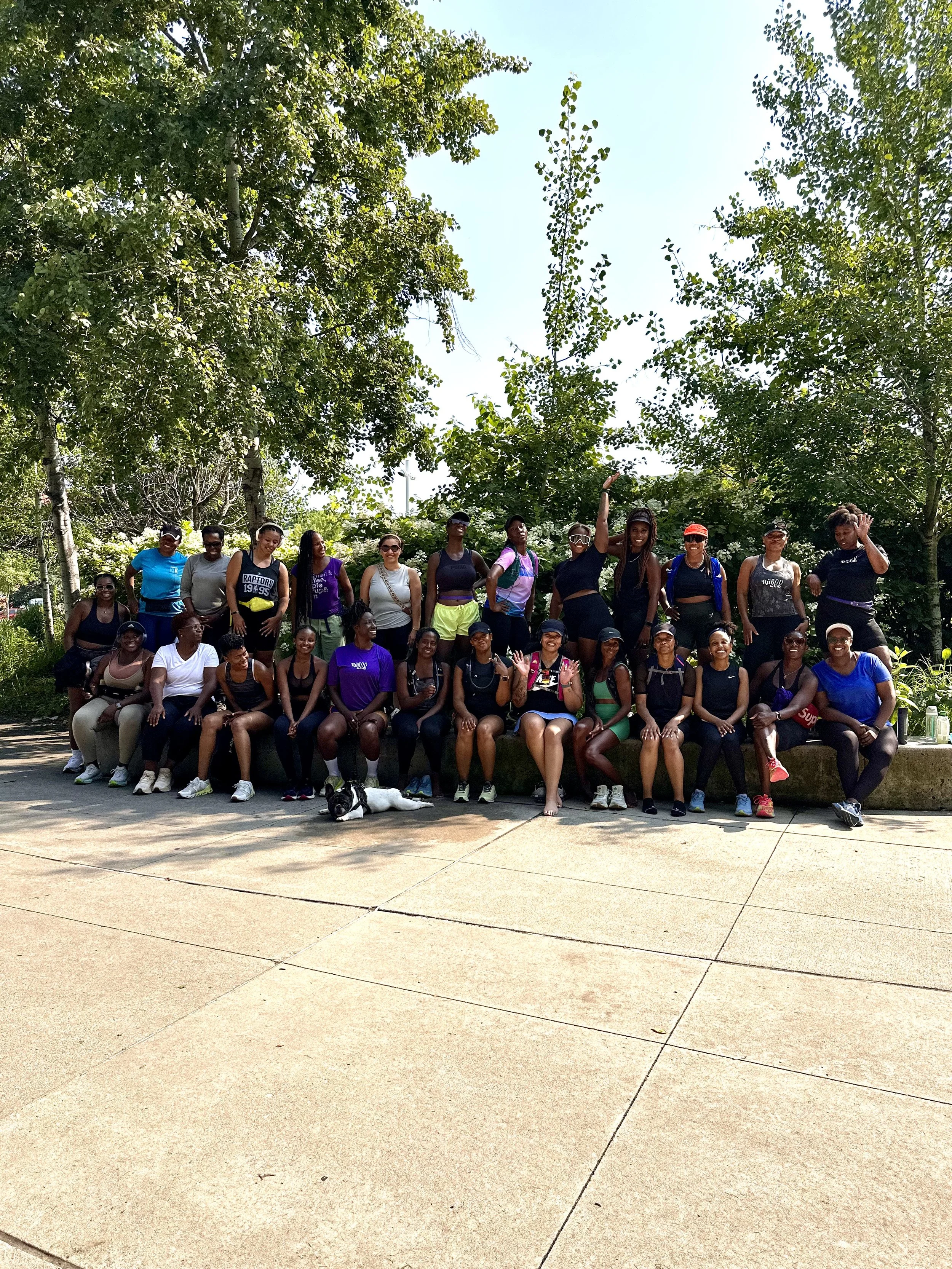 A large group of women in athletic clothing gathered outdoors under green trees, posing for a group photo with some members sitting on a ledge and others standing behind, with a dog lying on the ground in front.