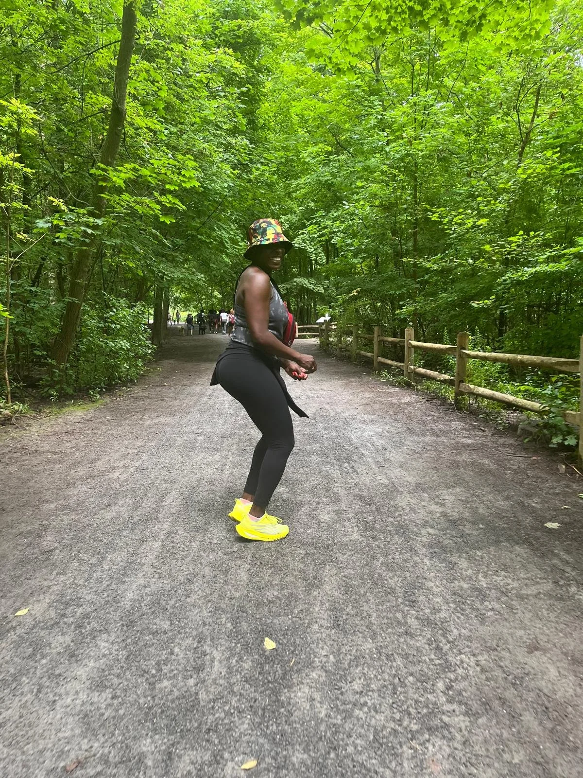 A woman wearing a colorful bucket hat, gray tank top, black leggings, and bright yellow running shoes, dancing on a forest trail with lush green trees around her.