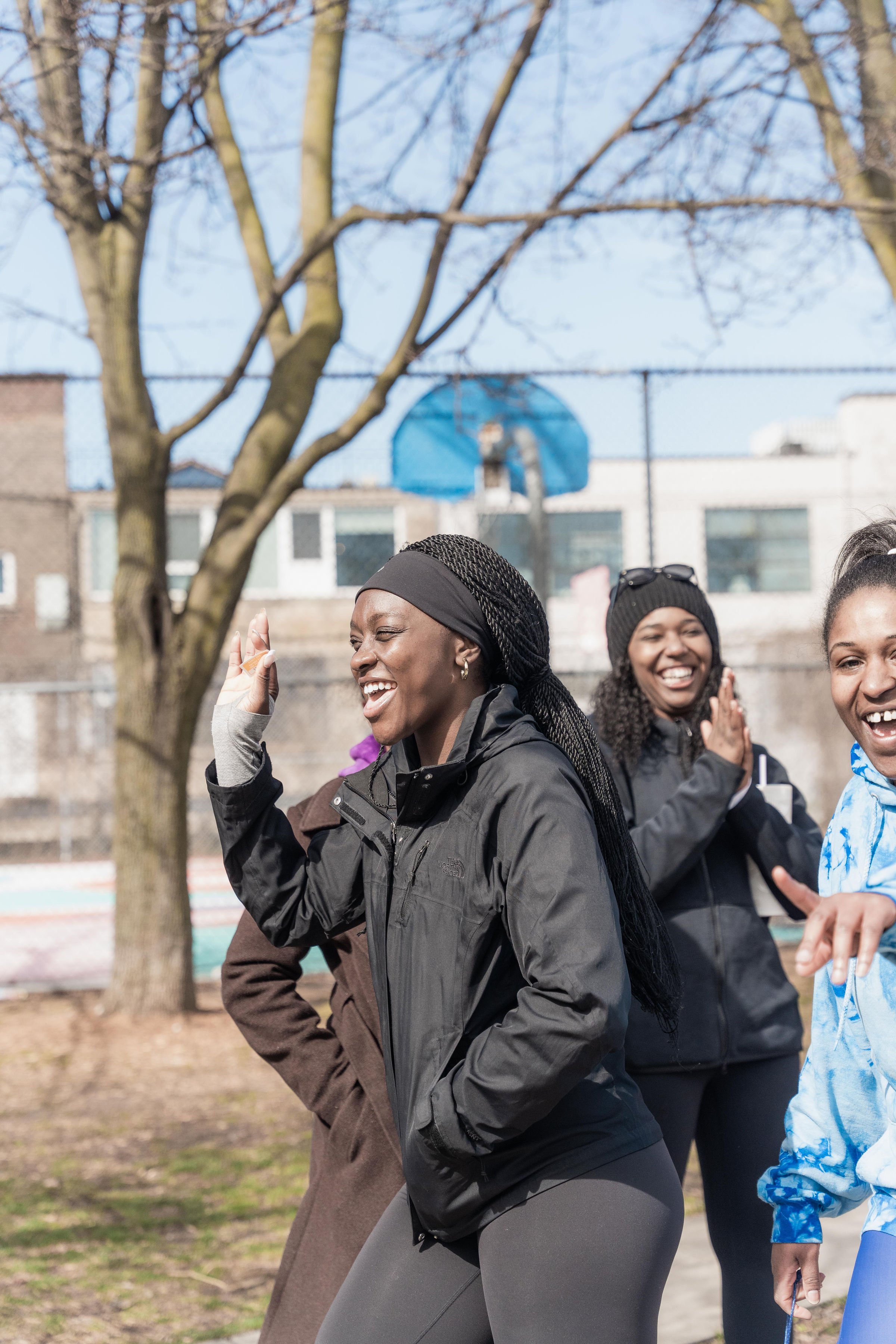 Group of women smiling and laughing outdoors on a sunny day near a tree and a basketball court.