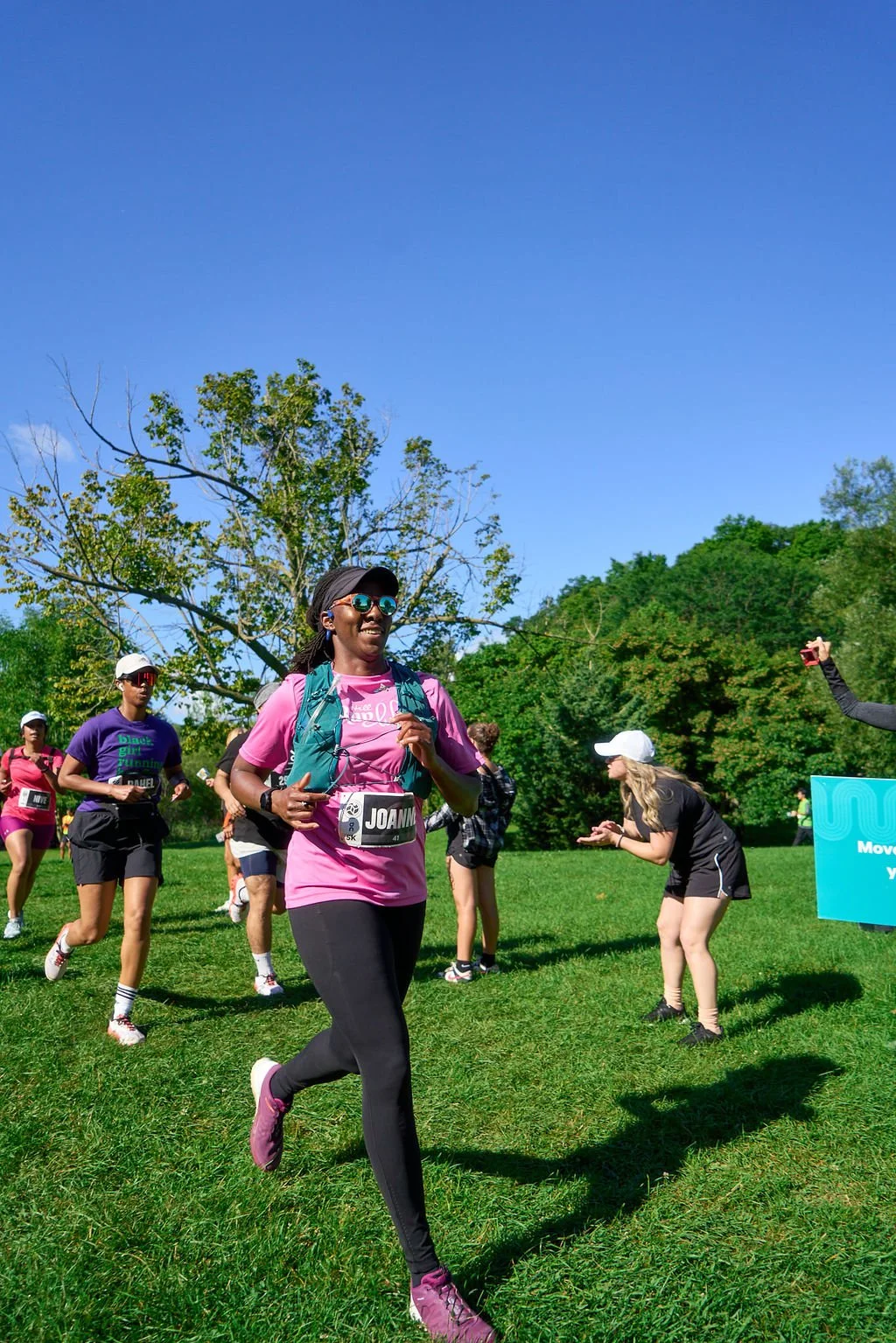 A group of runners participating in a race outdoors on a sunny day, with a smiling woman in pink running in the foreground and other runners and volunteers in the background, surrounded by green trees and a clear blue sky.