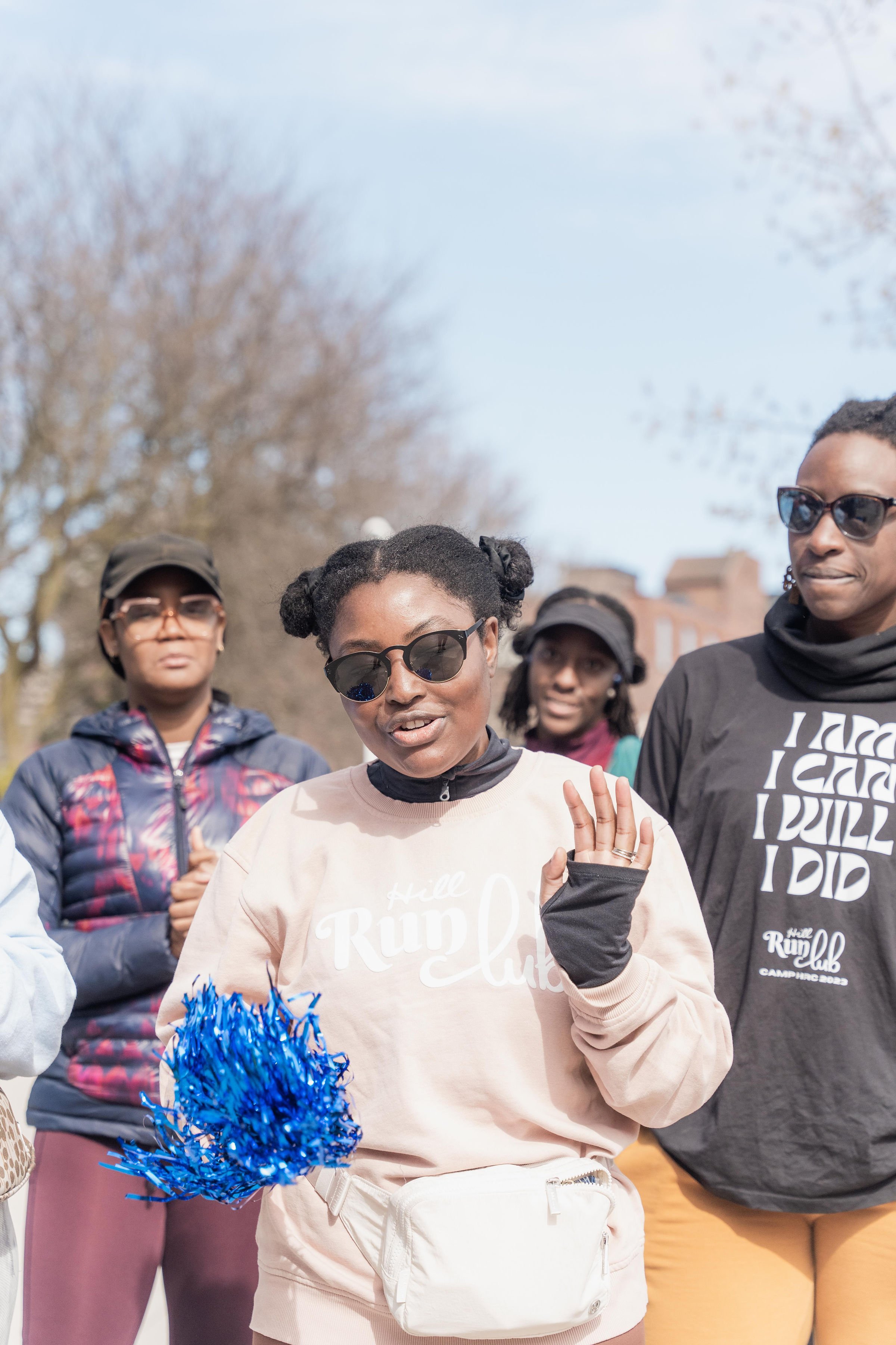 Group of women standing outdoors on a sunny day, wearing sunglasses and casual clothing, one woman is holding blue pom-pom, and they seem to be participating in an outdoor event or gathering.