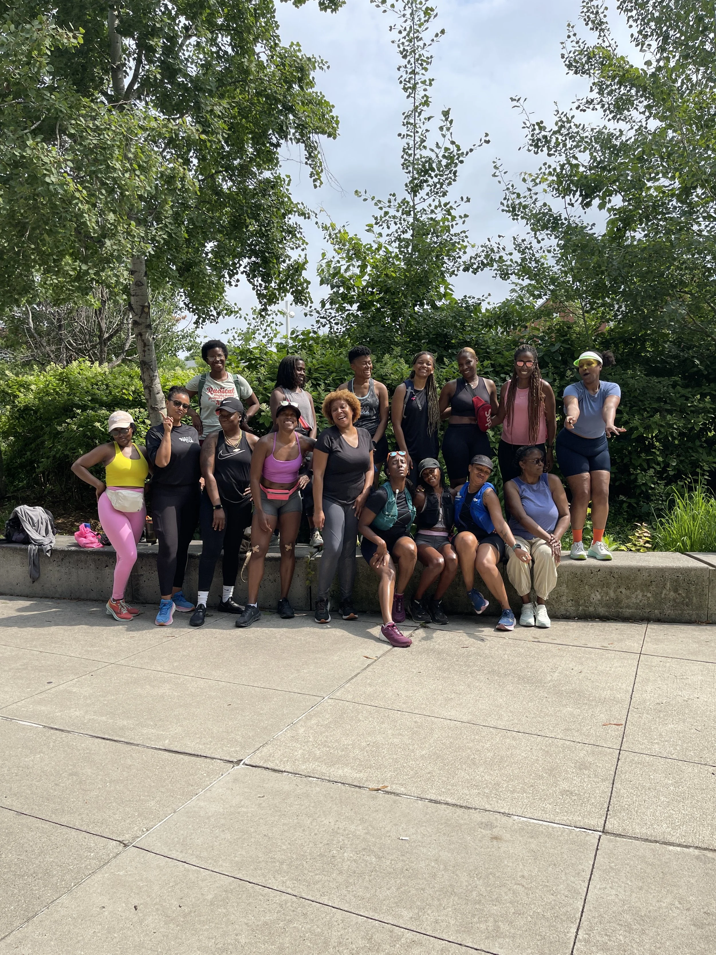 Group of women outdoors on a sidewalk, posing for a photo with greenery and trees in the background.