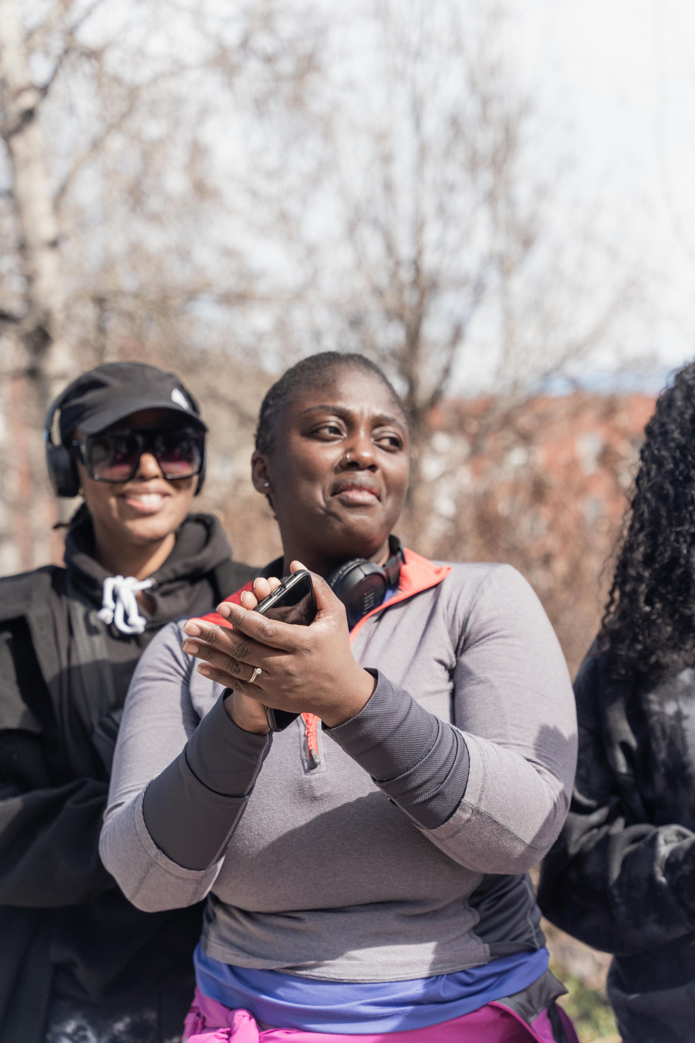 A woman holds a phone, looking emotional with a tearful expression, while two other women stand nearby outdoors in a park with bare trees in the background.