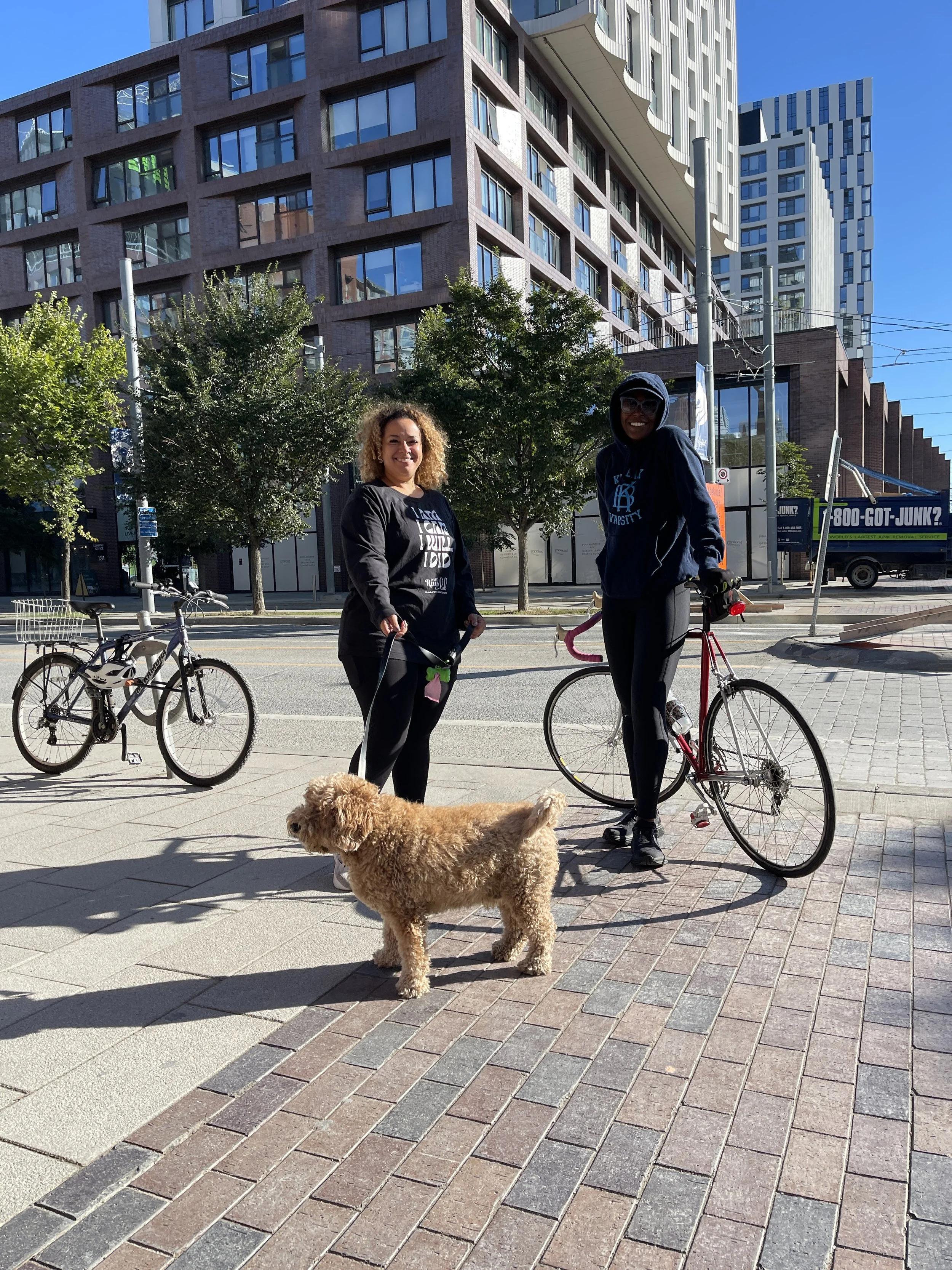 Two women standing on a city sidewalk with bicycles and a small dog, smiling at the camera. Behind them are trees, tall modern buildings, and a truck.