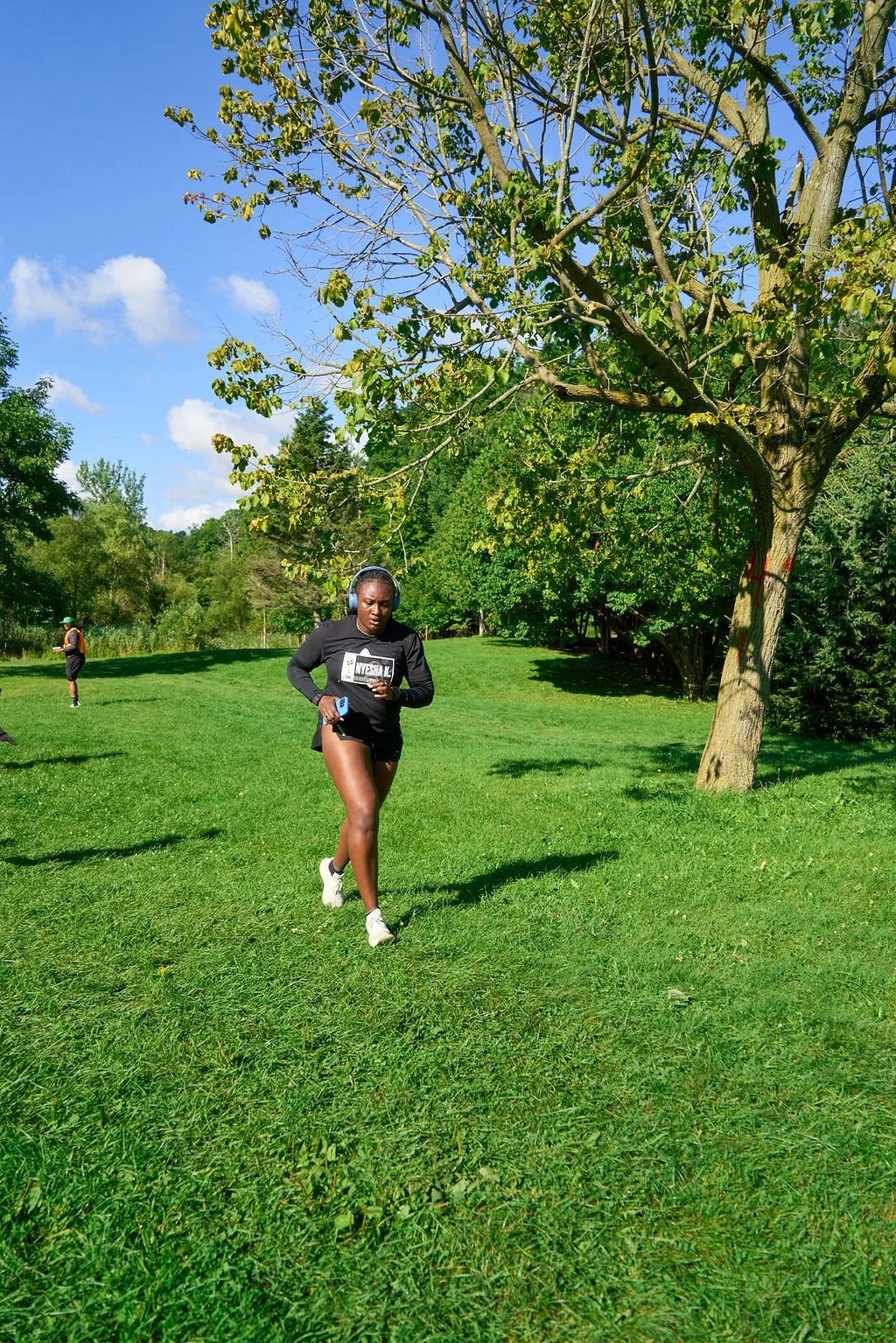 A woman running outdoors in a park on a sunny day, wearing headphones, a black sweatshirt, black shorts, and white sneakers. There are other runners in the background, and the park has lush green grass, trees, and a clear blue sky.