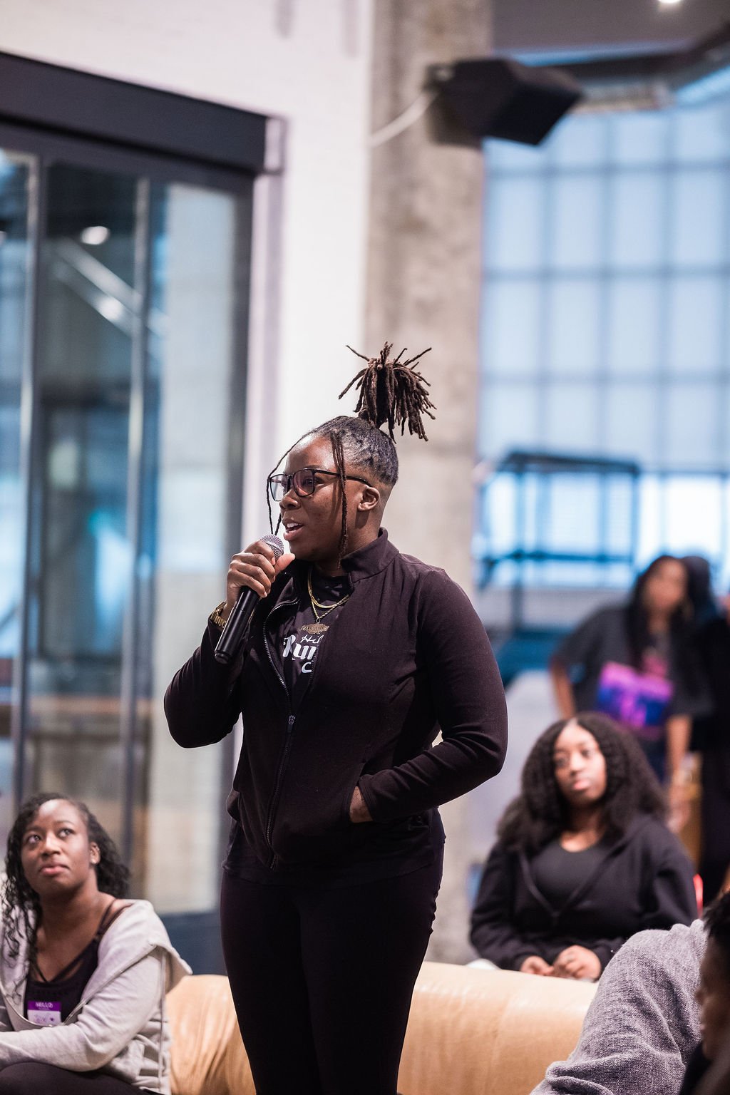 A woman with dreadlocks tied up in a high ponytail, wearing glasses, holding a microphone, speaking to an audience in an indoor setting.