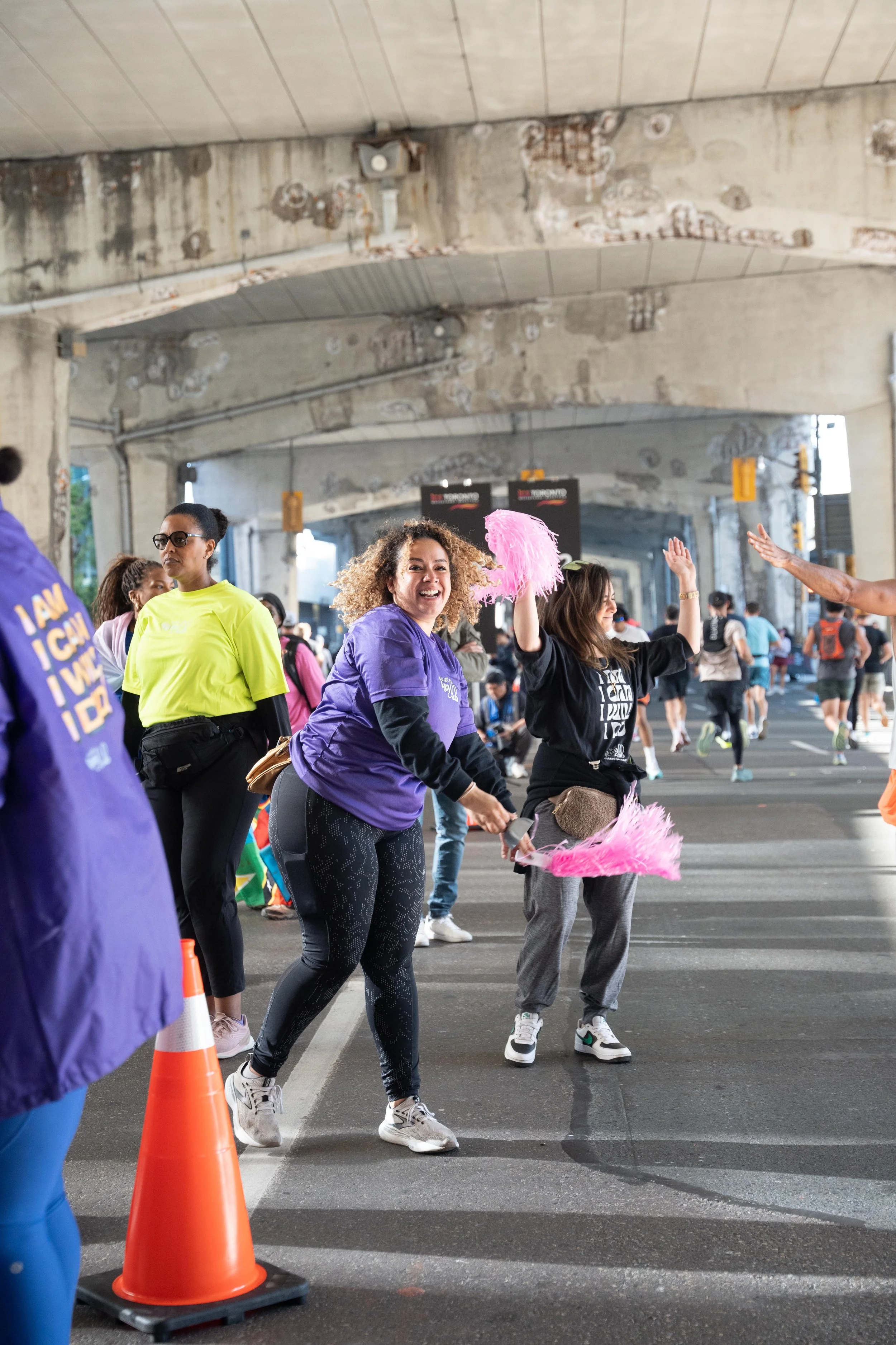 People participating in a walking or running event under an overpass, with some smiling and waving, and others walking or standing.