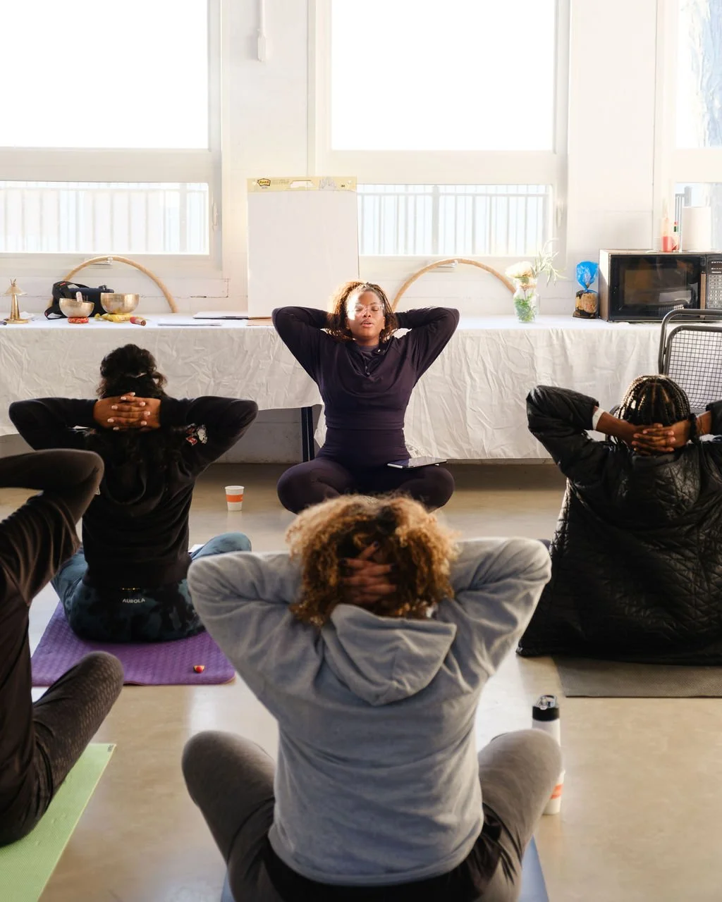 A group of people participating in a yoga or meditation session in a bright room, seated on yoga mats with their hands behind their heads, led by a woman sitting cross-legged at the front.