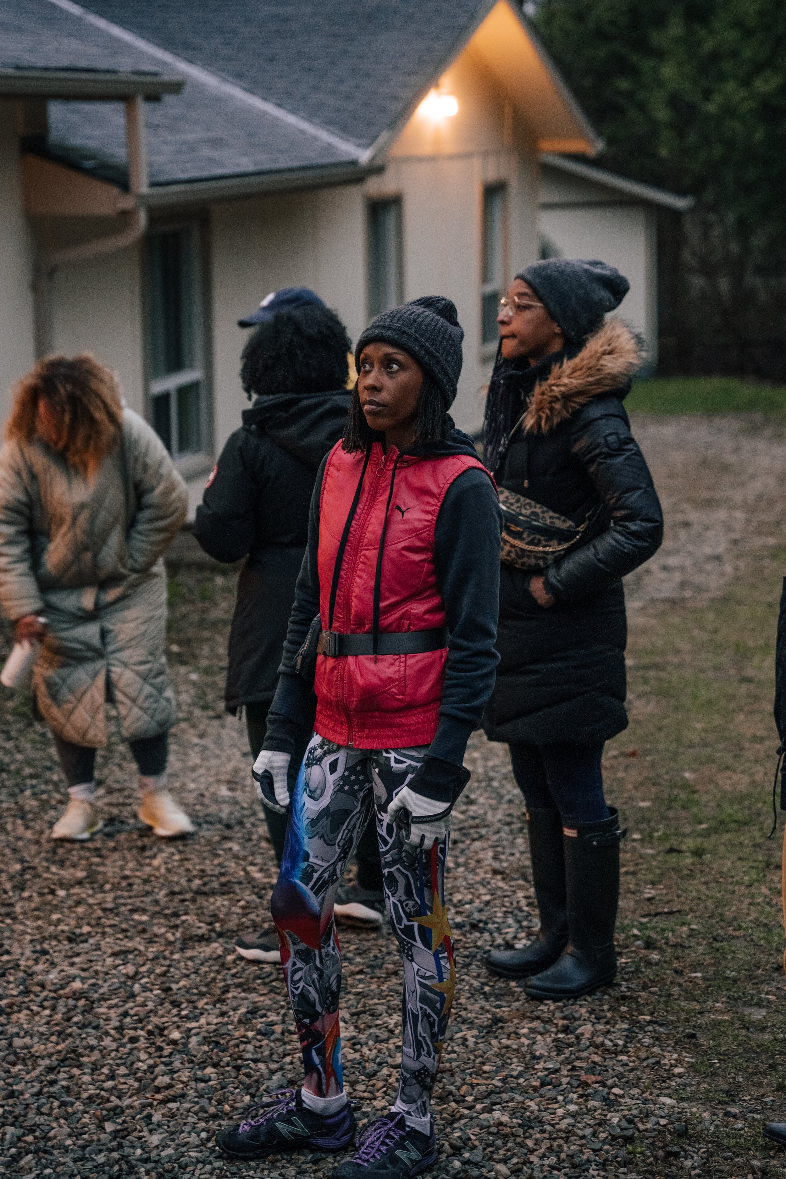 A group of women standing outside near a building during twilight, dressed in warm clothing including jackets and beanies, with one woman in the foreground wearing colorful leggings and a red jacket.