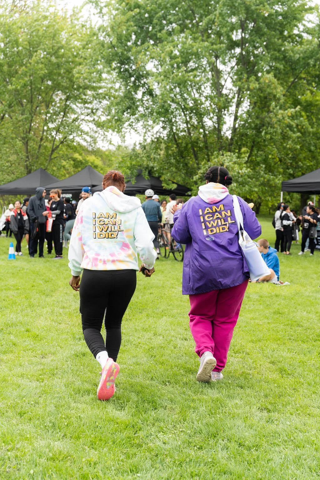Two women walking toward a gathering with tents in a park, wearing hoodies that say 'I AM I CAN I WILL I DID' on the back.