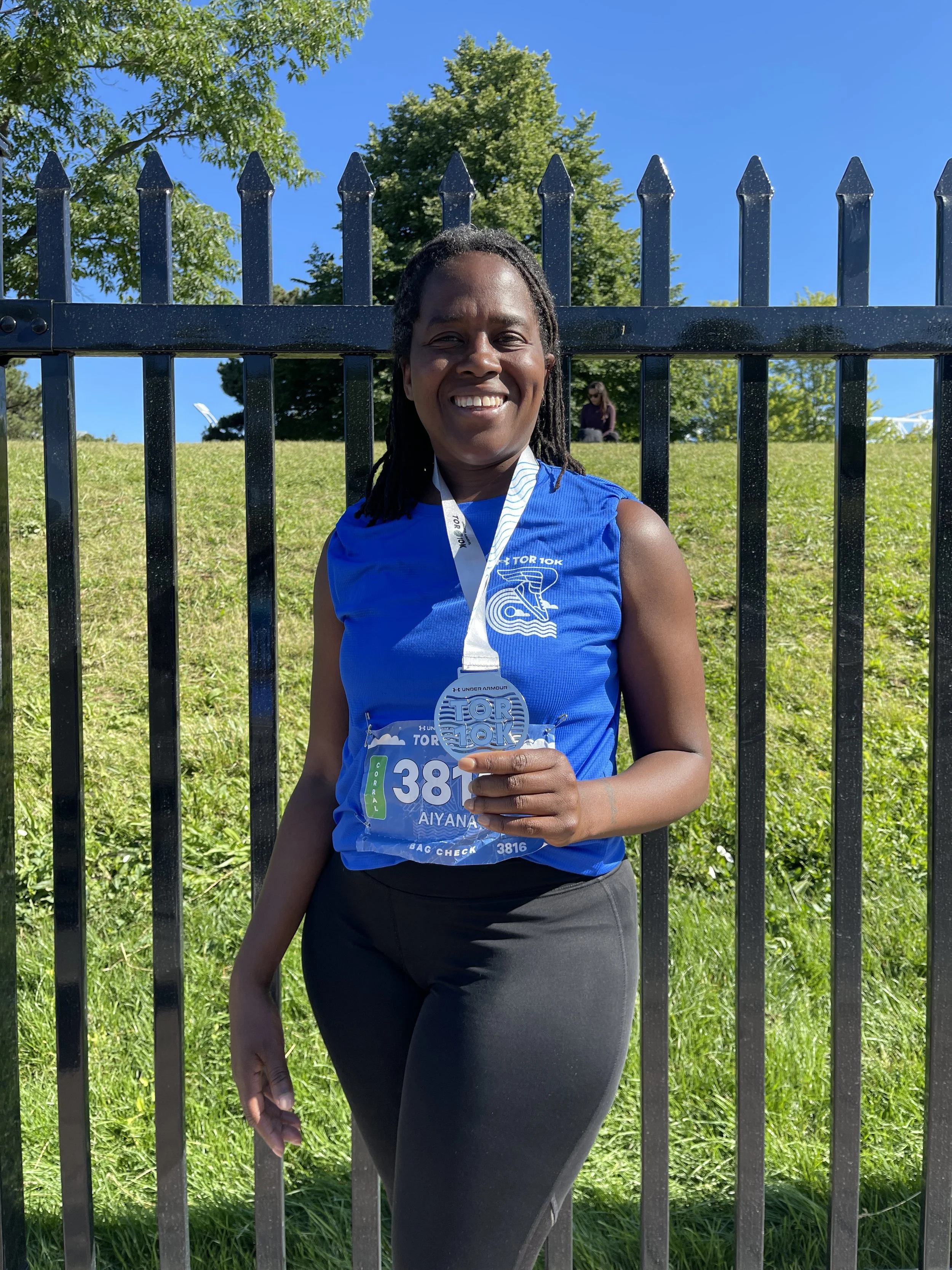 Woman smiling and holding a race medal after finishing a race, standing in front of a black metal fence with green grass and trees behind her.