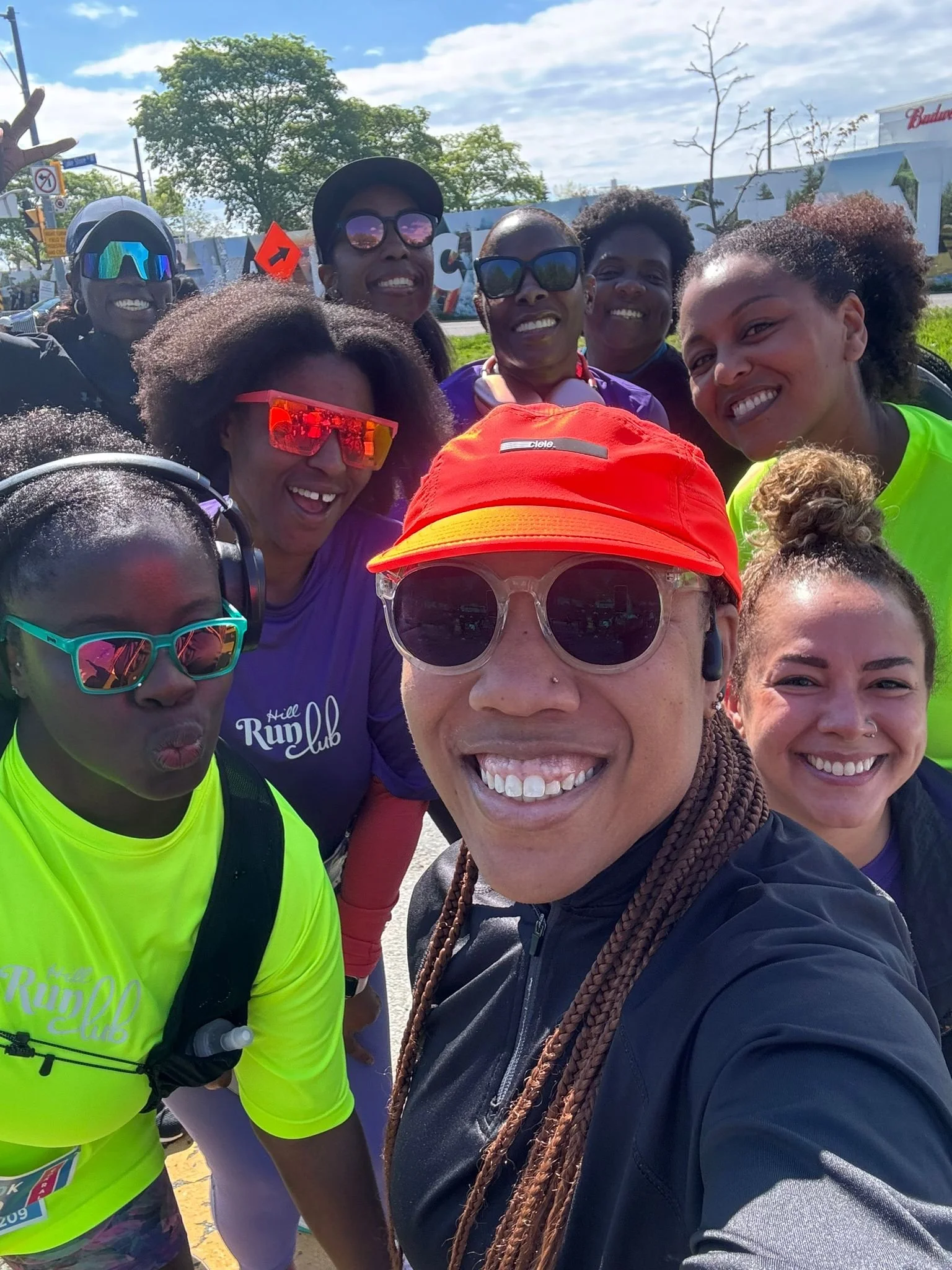 Group of smiling women, some wearing bright athletic shirts, sunglasses, and hats, taking a selfie outdoors on a sunny day with trees and a building in the background.