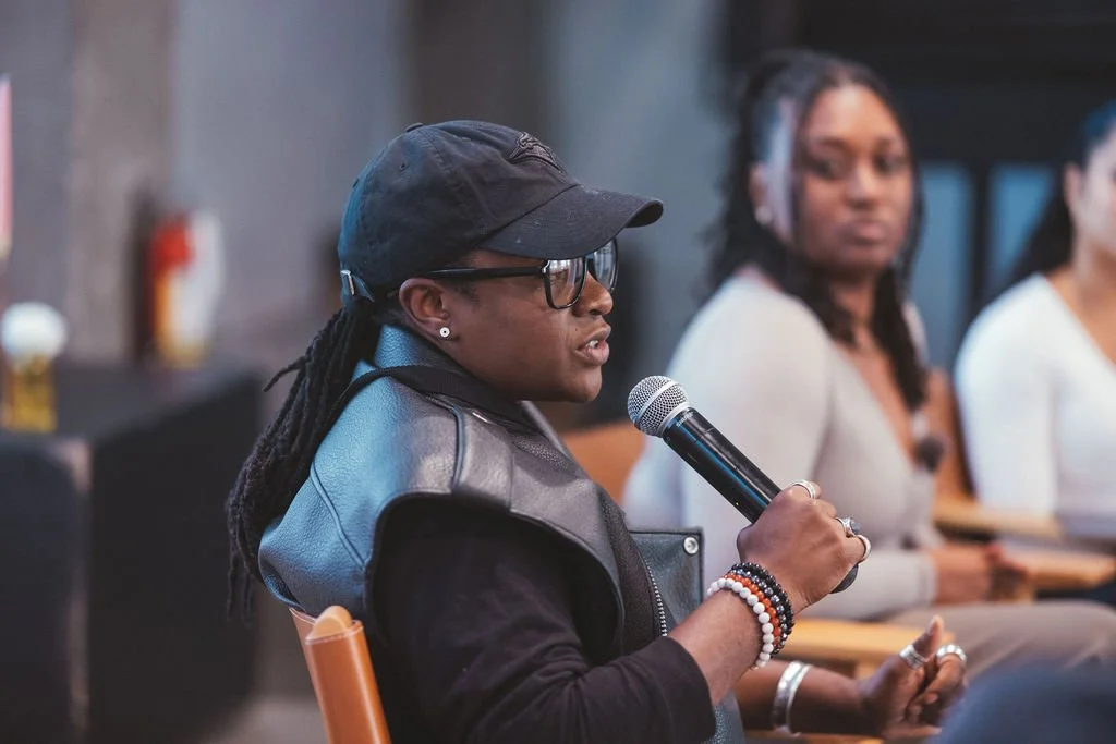 A woman with braided hair, glasses, and earrings speaking into a microphone during a panel discussion, with two women sitting behind her.