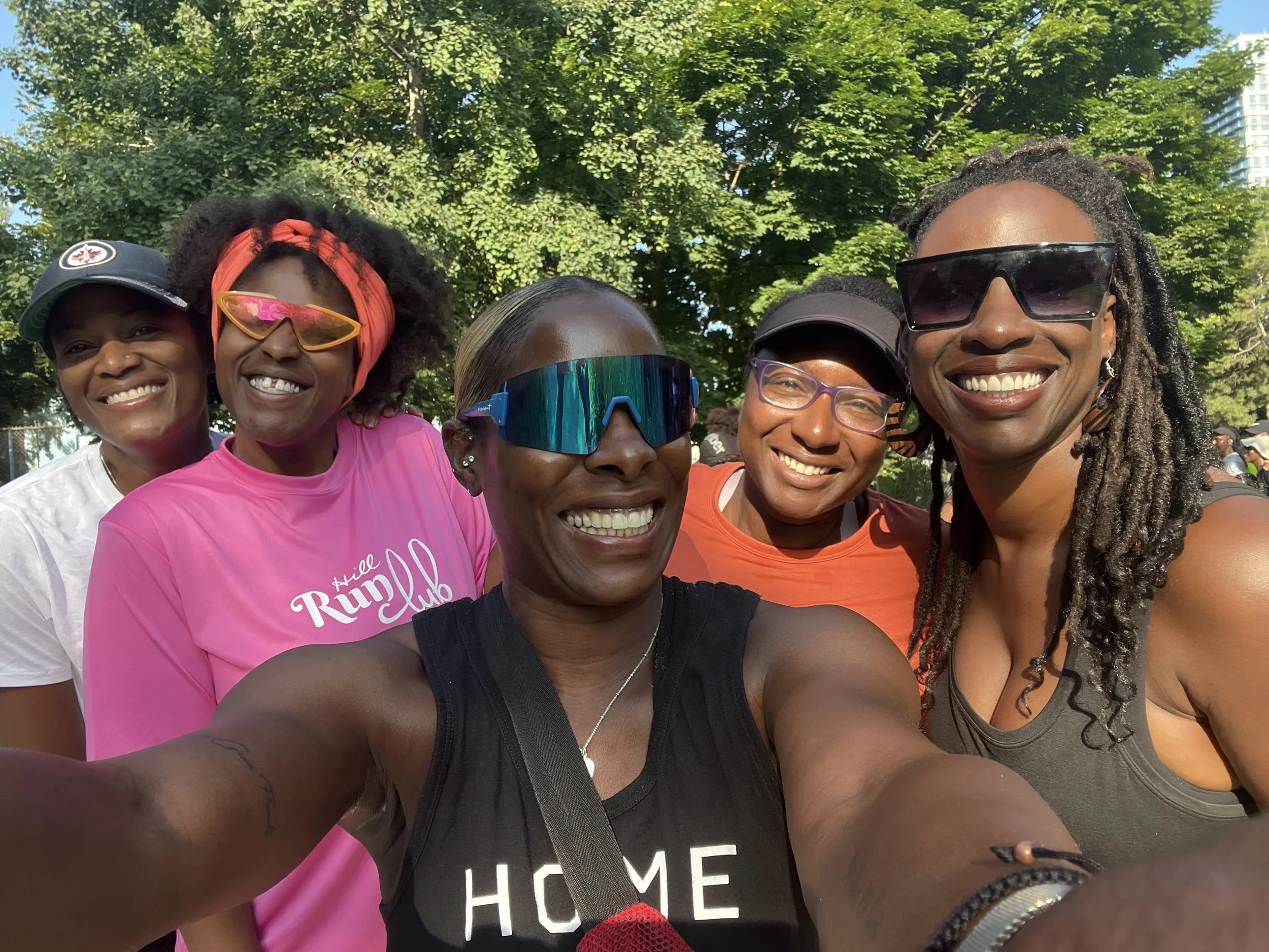Group of five smiling women outdoors in a park, wearing athletic clothing and sunglasses, with green trees and a building in the background.