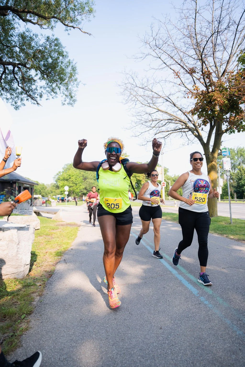 Group of women participating in a 5K race, smiling and running on a park trail in warm weather, with trees and a clear sky in the background.