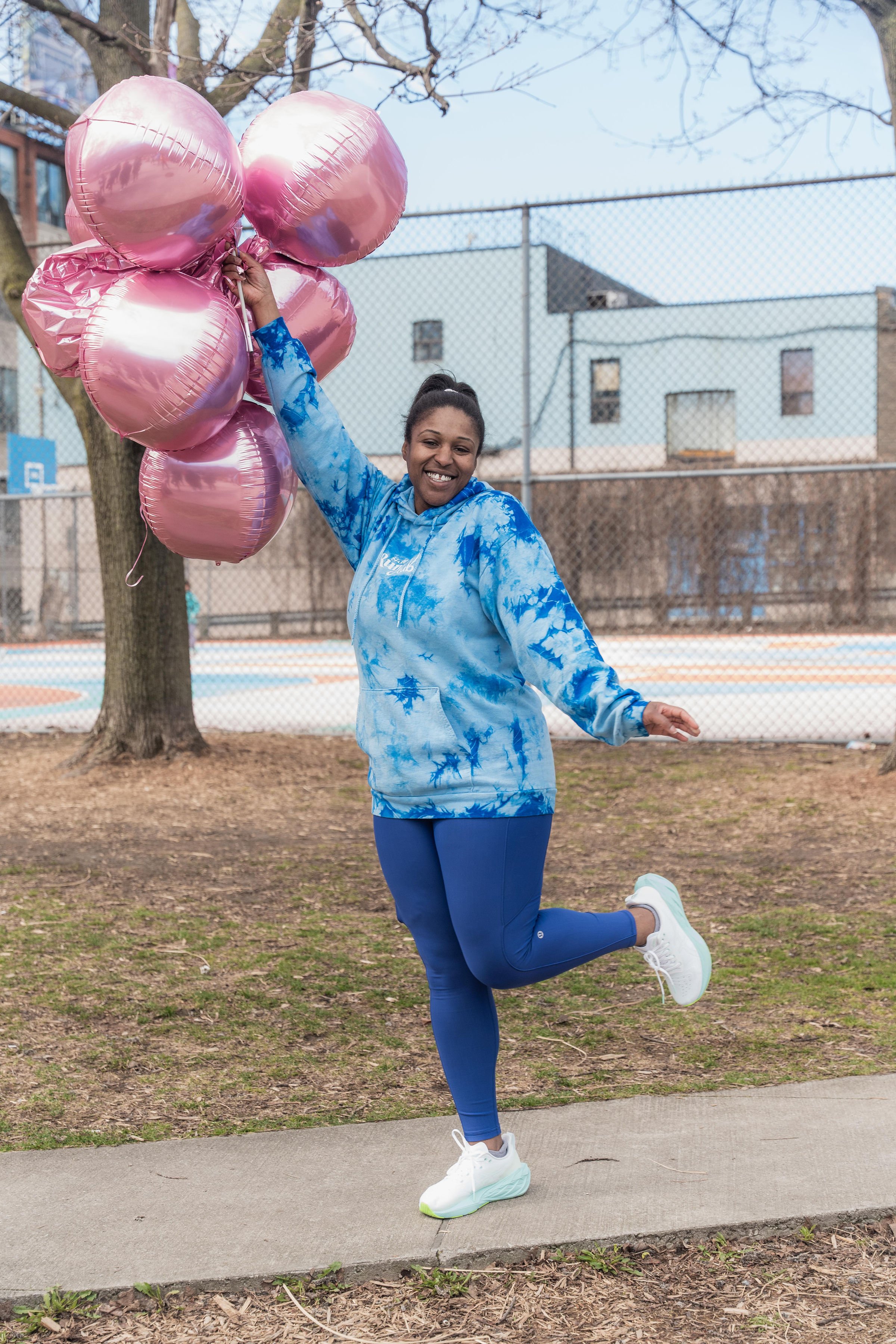 Woman smiling, wearing a blue tie-dye hoodie and blue leggings, holding a bunch of pink balloons while standing on a sidewalk in a park.