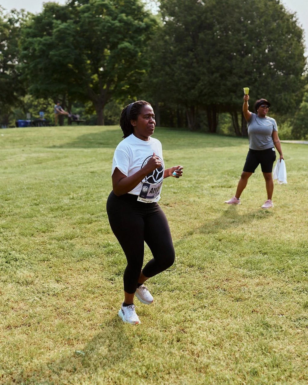 A woman runs on a grassy field during a park or outdoor event, with trees and another woman with a water bottle and towel in the background.