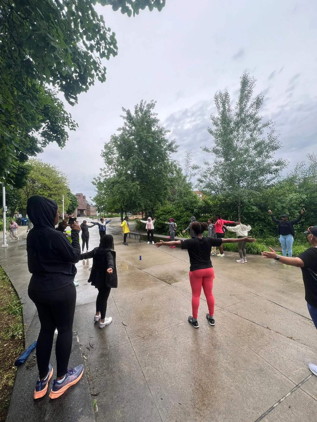 A group of people participating in an outdoor exercise class in a park, standing with arms extended to the sides.