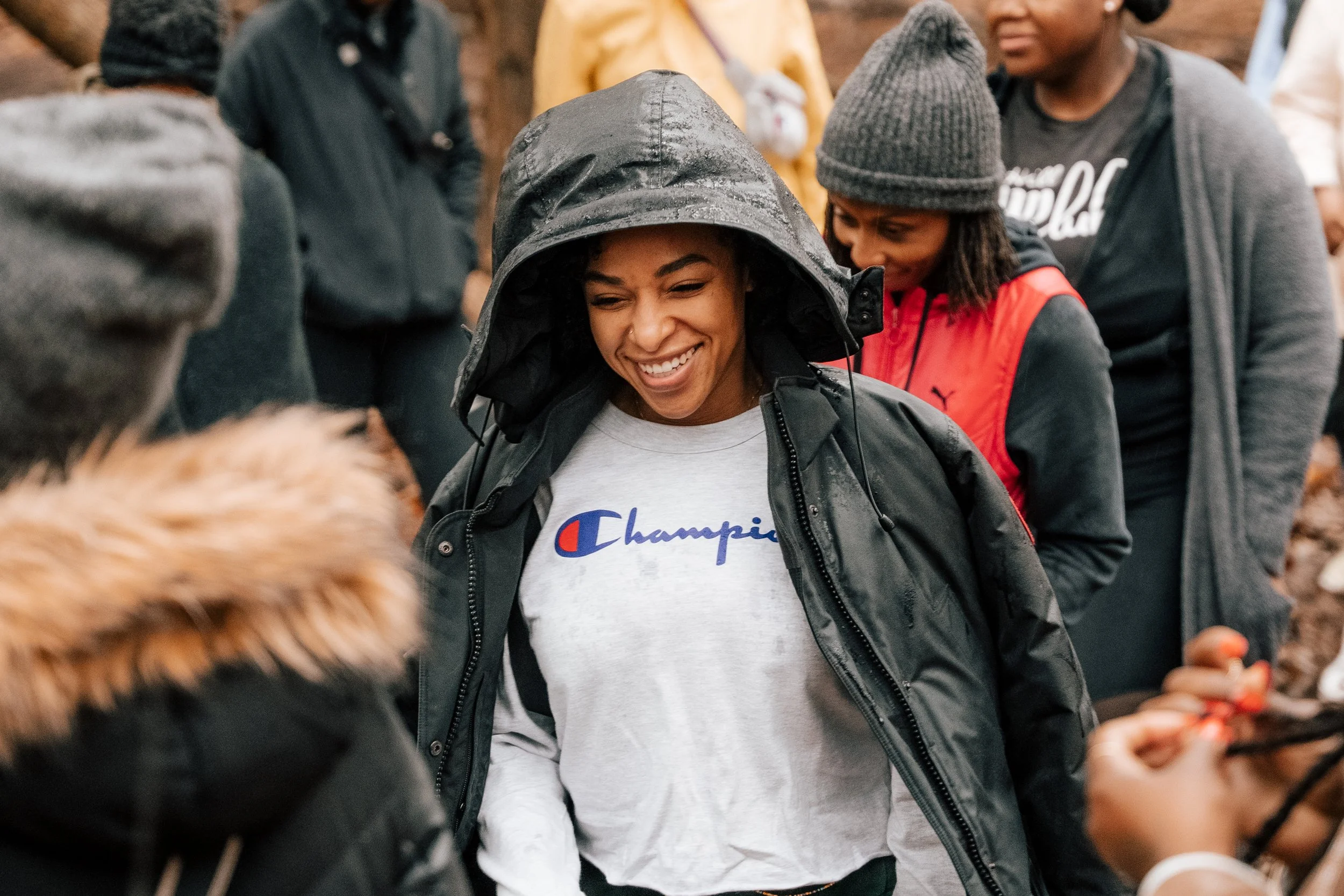 A group of people outdoors, with one woman in the center smiling, wearing a black rain jacket with the hood up and a gray shirt with a 'Champion' logo. Others around her are dressed warmly, some with hats and jackets, and they appear to be engaged in