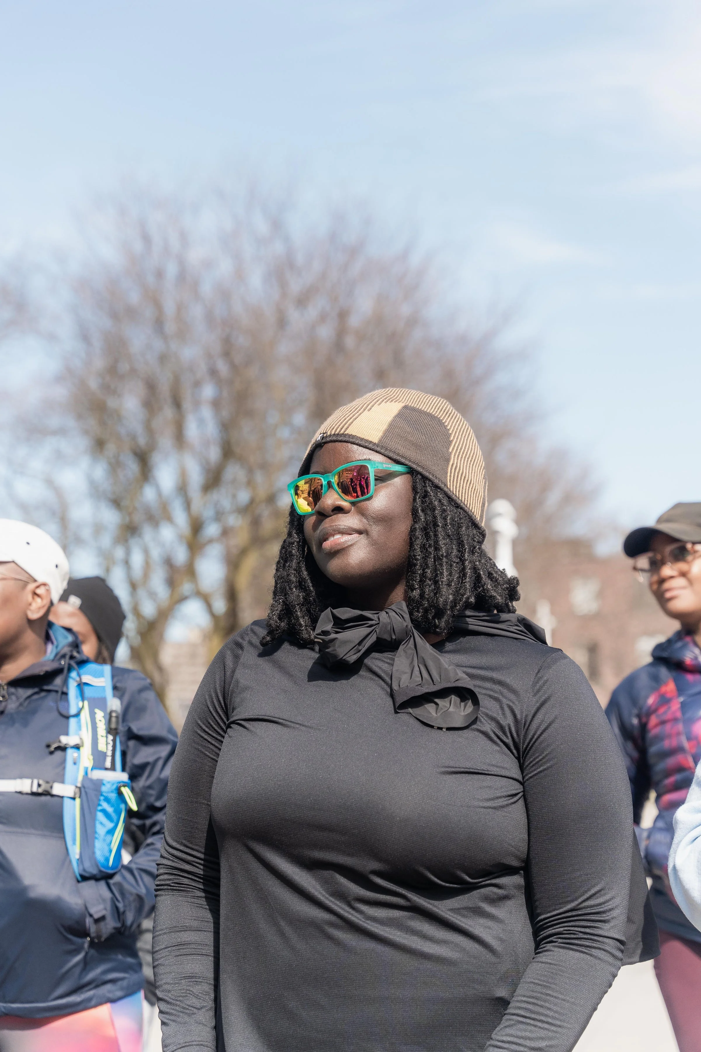 A woman wearing sunglasses, a beanie, and a black long-sleeve shirt, standing outdoors among other people with trees and a clear sky in the background.