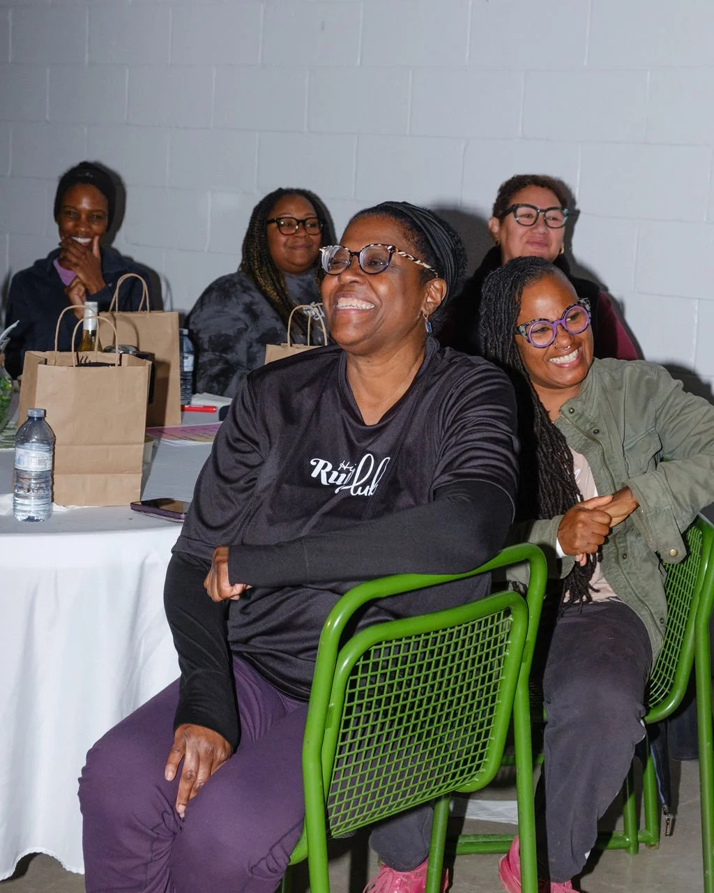 A group of women smiling and laughing at a seated event, with tables, brown paper bags, water bottles, and a gray brick wall in the background.