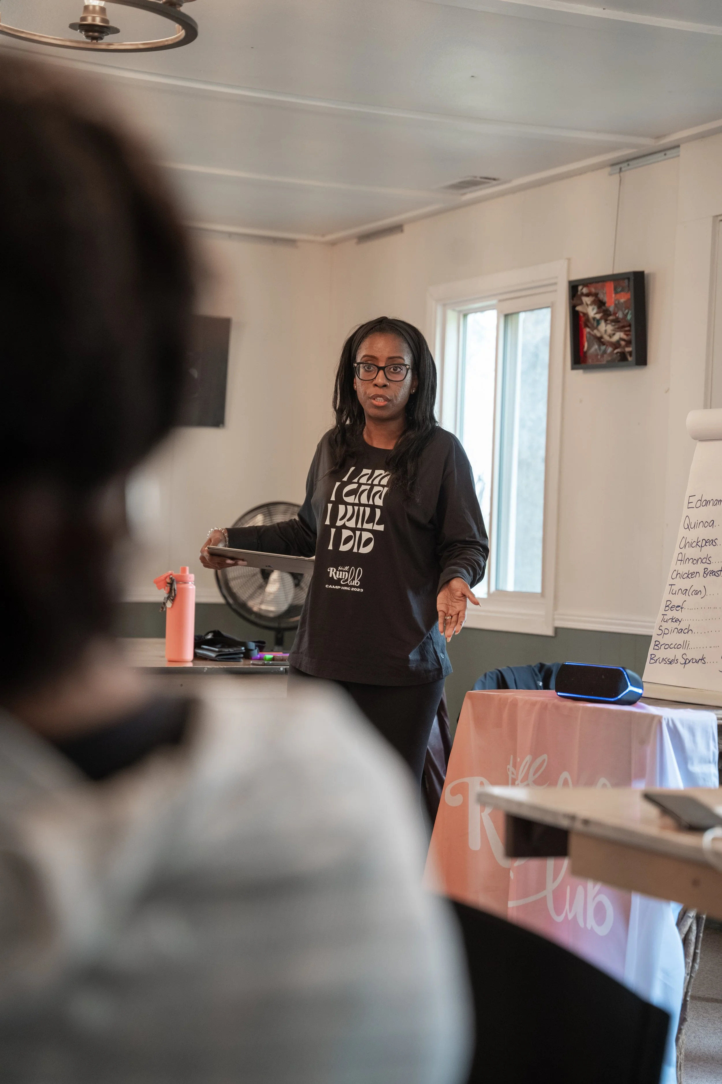 A woman standing in front of a room with windows, speaking to an audience during a presentation or meeting, with a pink tablecloth and a whiteboard visible in the background.