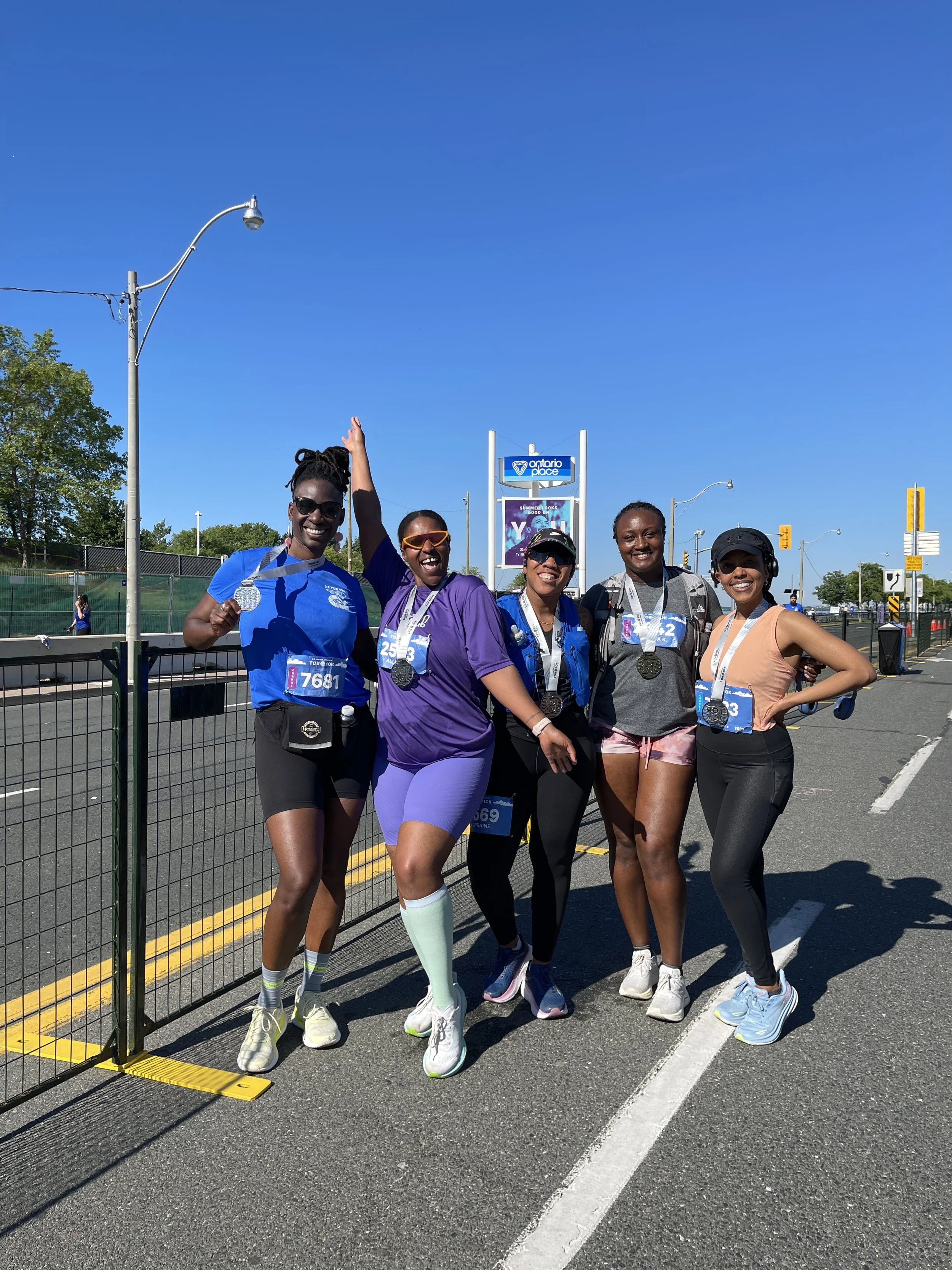 Five women celebrating after a race, wearing medals, race bibs, and athletic clothing, standing on a street near a fence with a clear blue sky overhead.